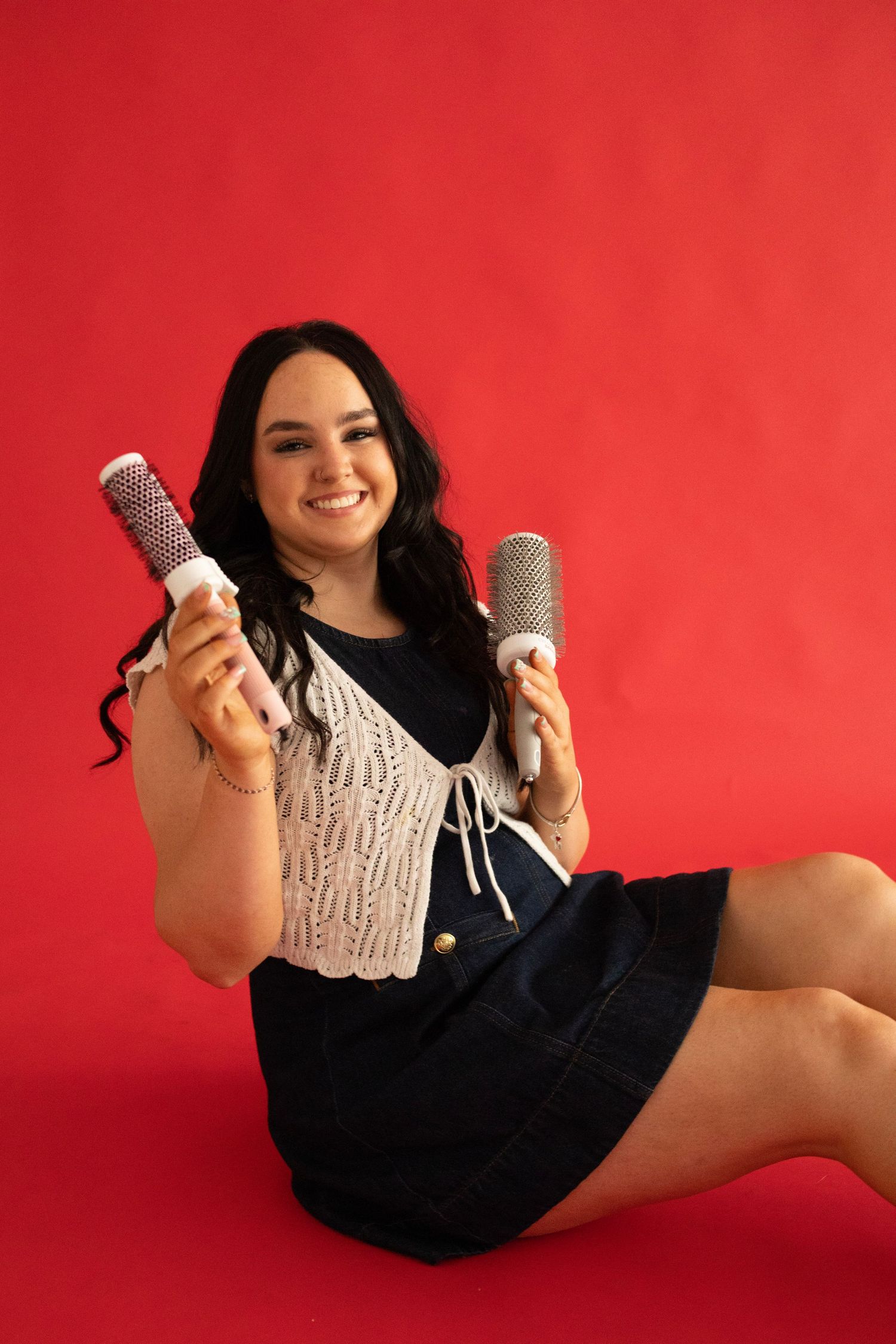 Young woman sitting on red background smiling while holding two round hair styling brushes in a denim skirt.