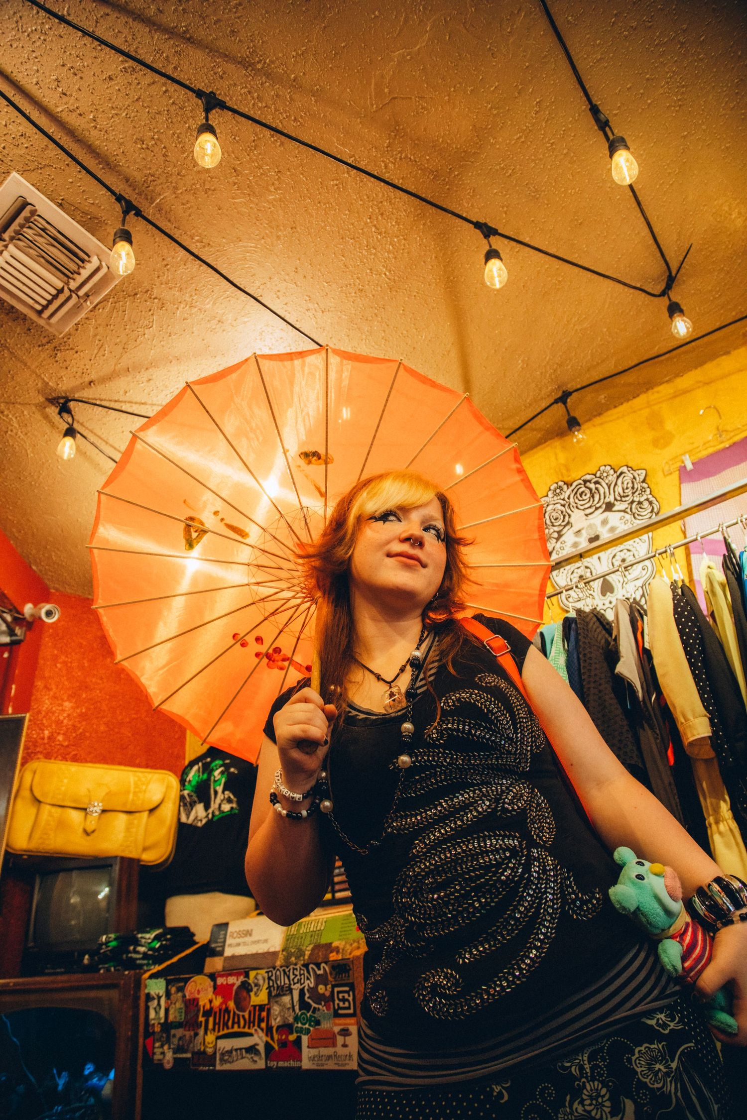 Red-haired woman holding orange parasol in colorful vintage shop with string lights overhead.