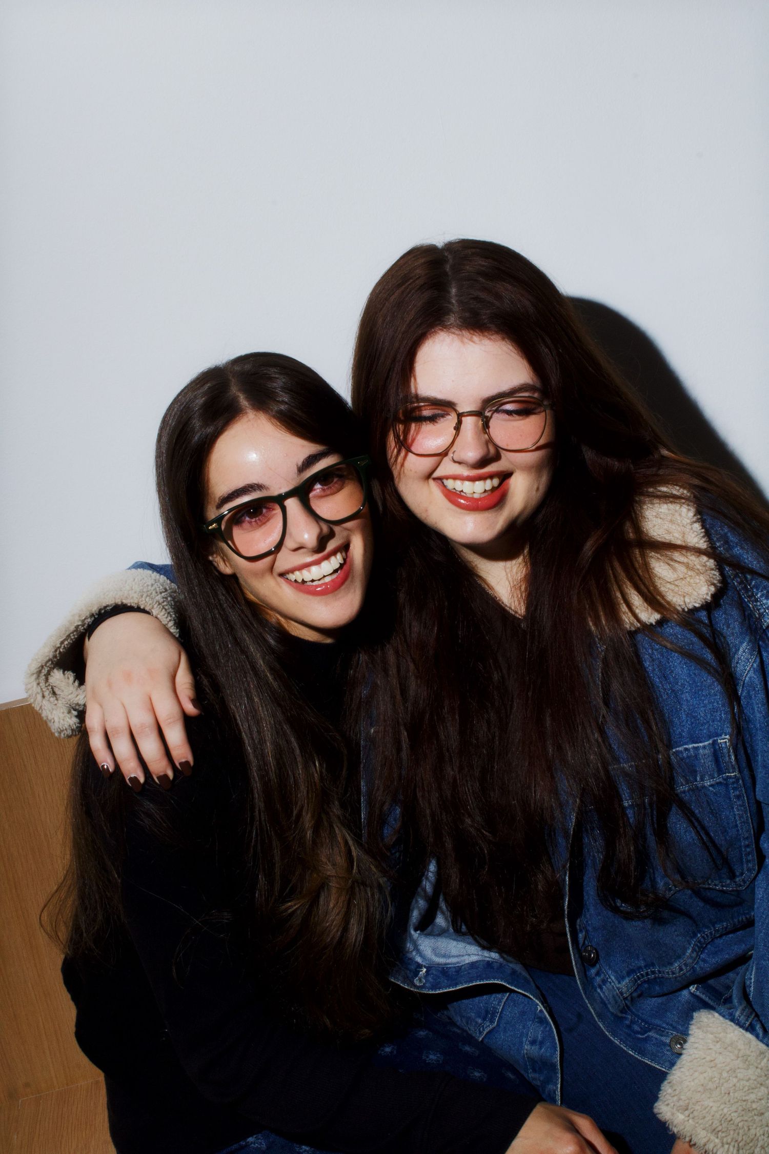 Two smiling young women with long dark hair wearing glasses embracing in a joyful portrait photo.