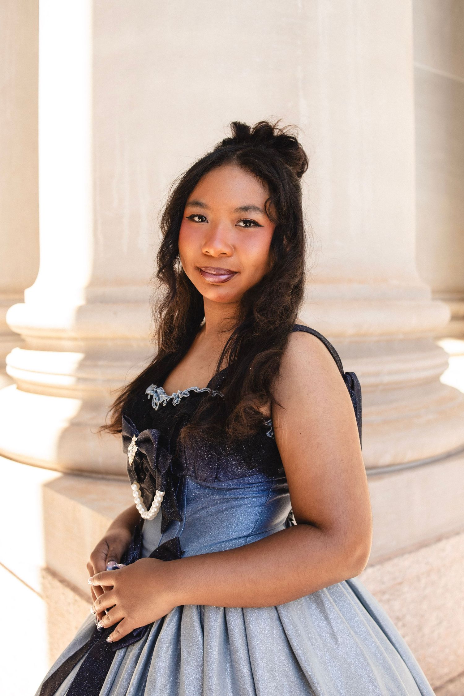 Young woman in elegant black and blue ball gown posing by stone columns in golden sunlight.
