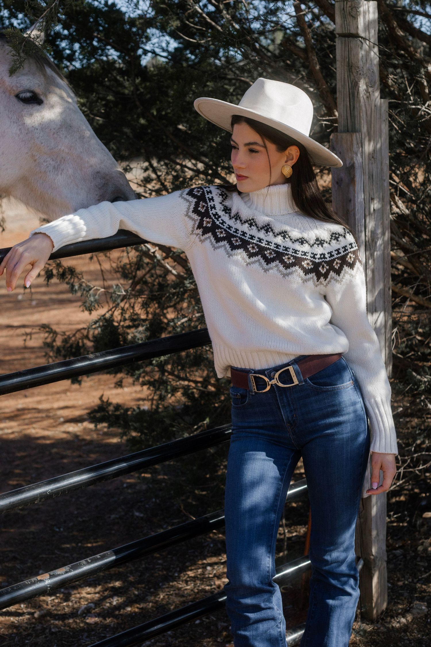 Woman in white Fair Isle sweater and jeans with hat petting white horse at ranch fence.