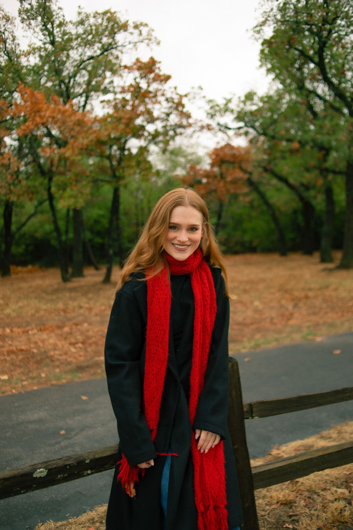 Young woman in black coat and red scarf smiling in autumn park with colorful foliage.