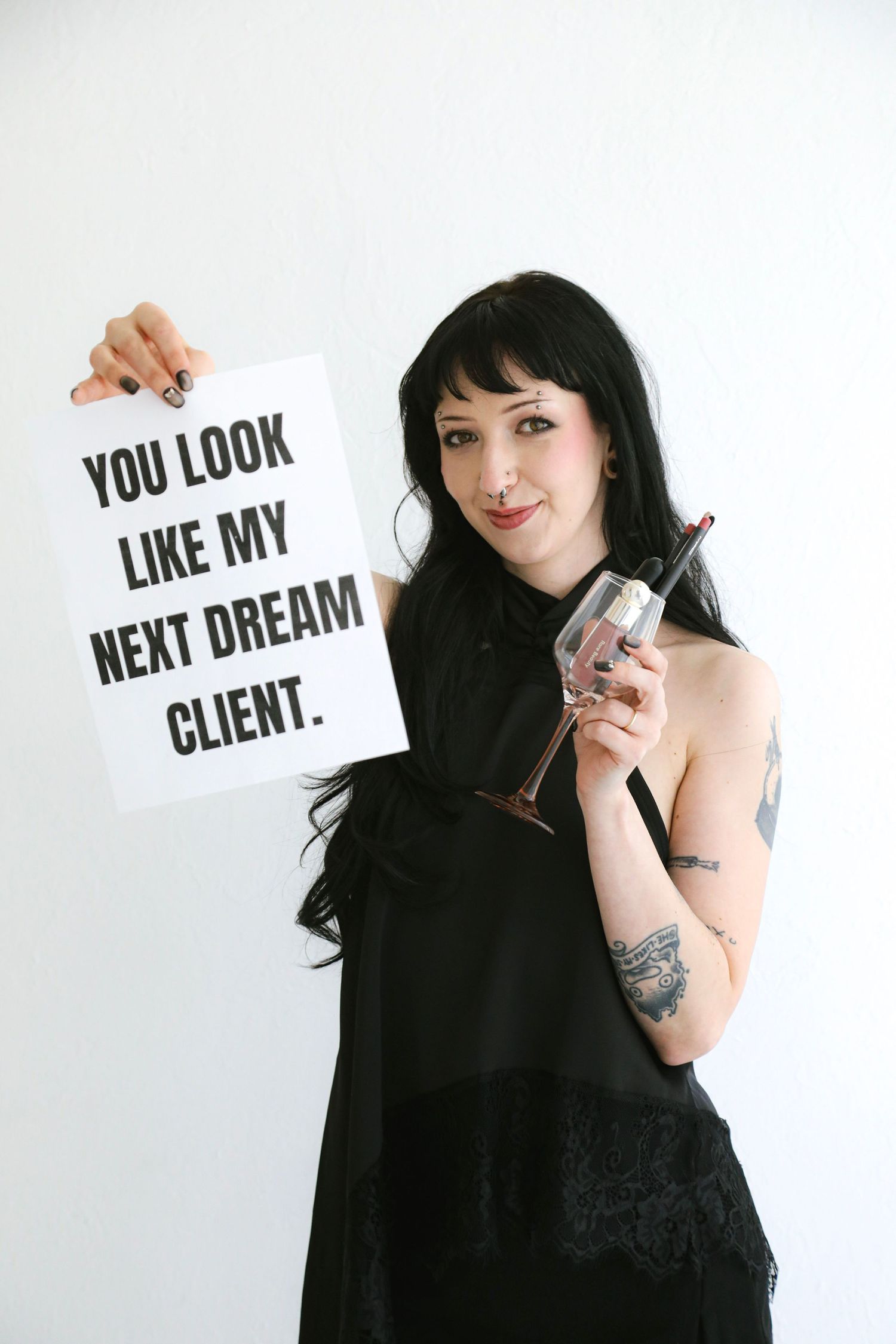 Woman in black dress holding a sign reading 'You Look Like My Next Dream Client' while holding a wine glass.