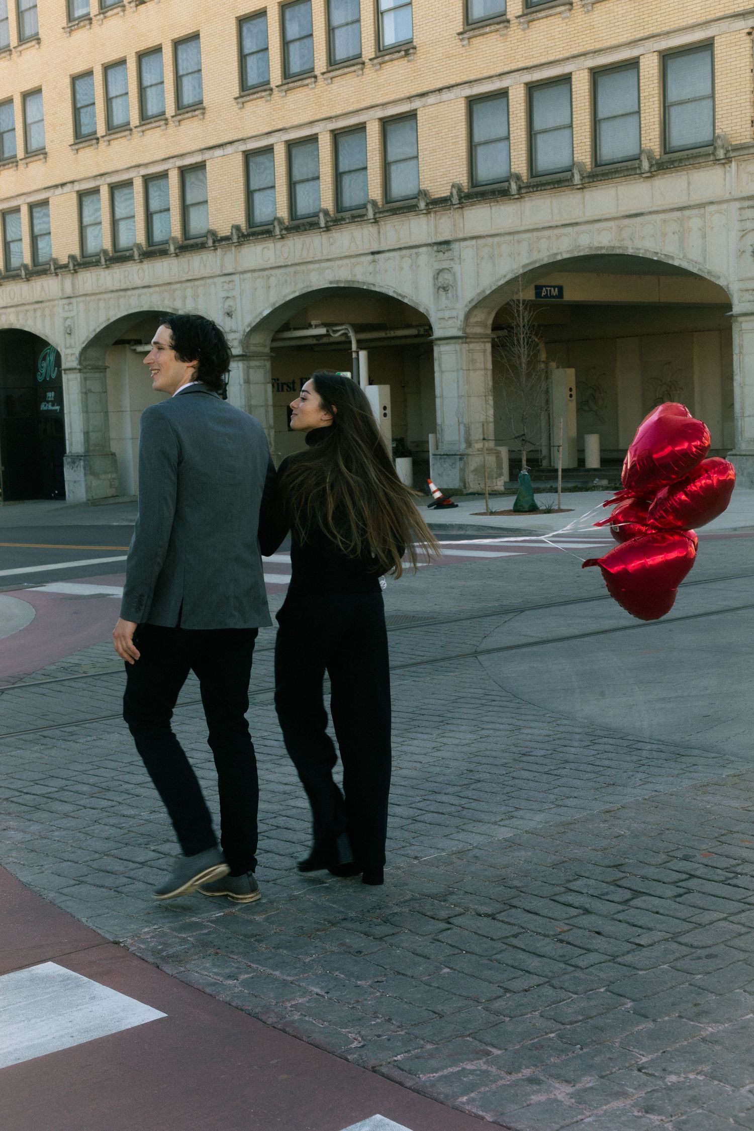 A couple walks together on a city street, the woman holding red heart-shaped balloons.