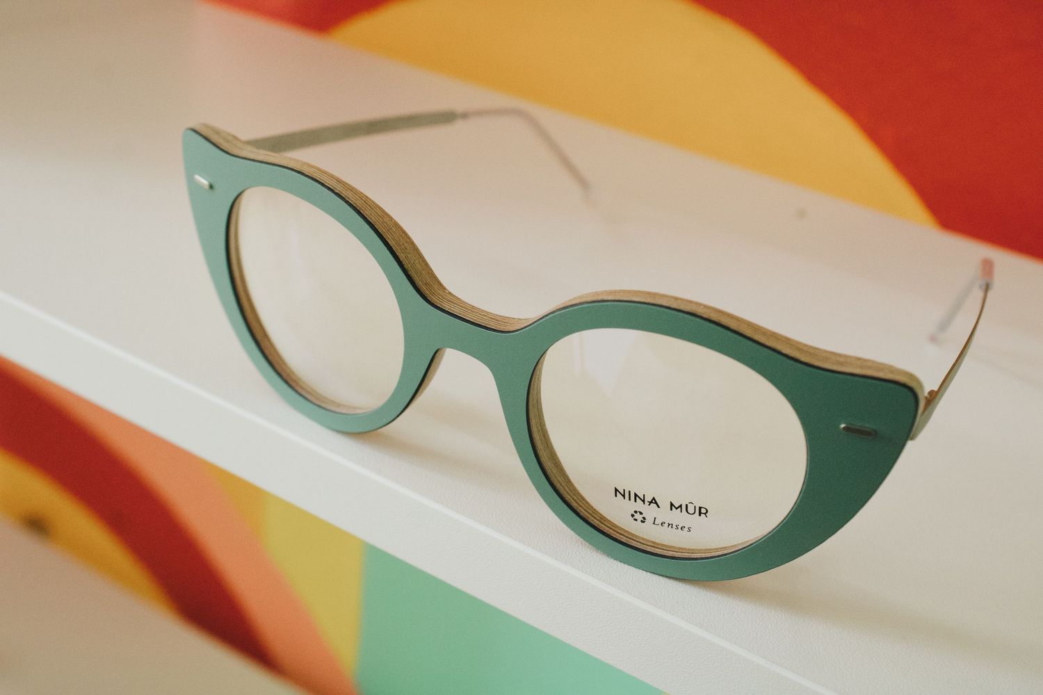 Teal and gold cat-eye Nina Mur eyeglass frames displayed on a white shelf in an optical boutique.
