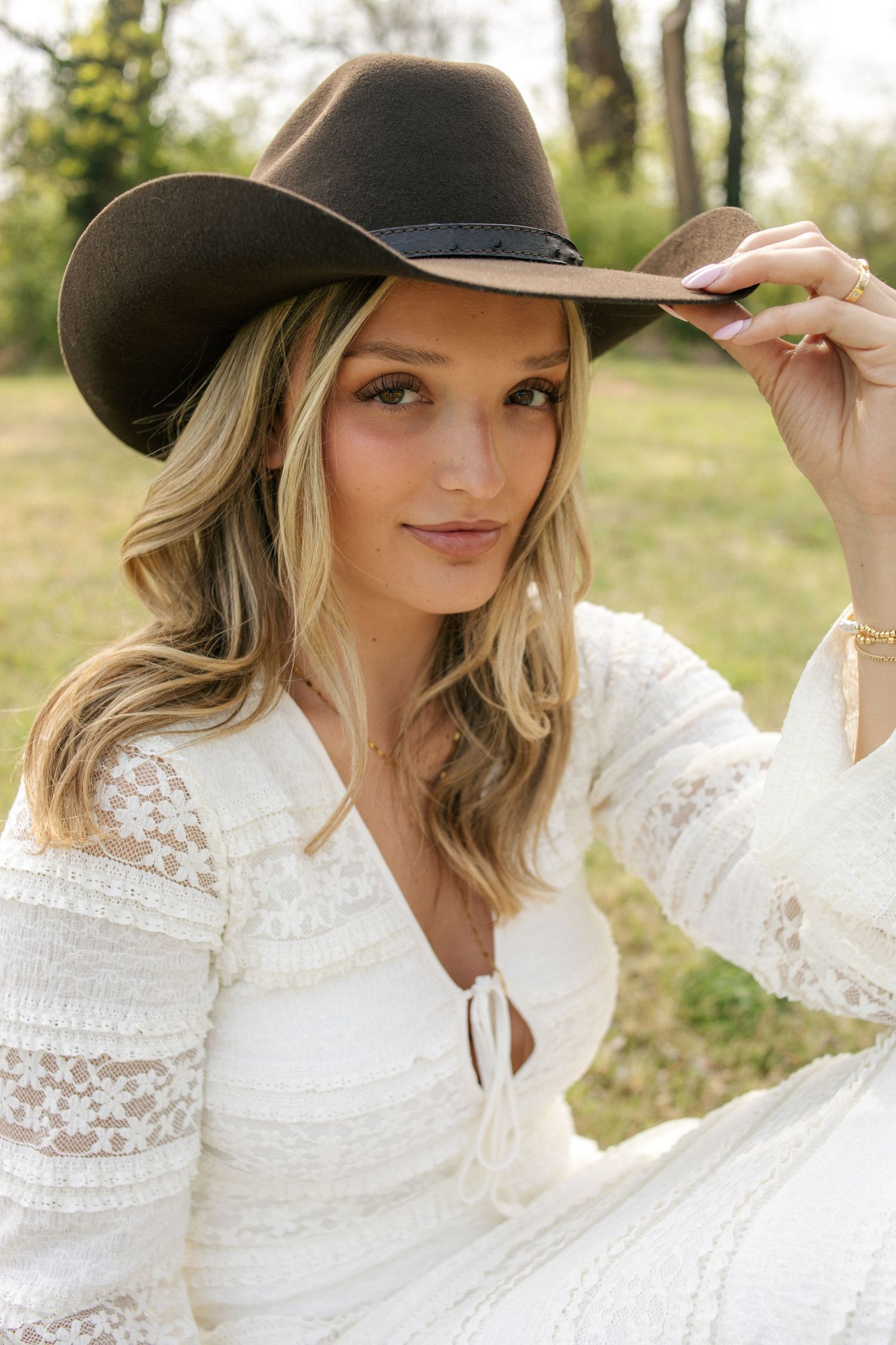 Blonde woman in white lace dress wearing brown cowboy hat outdoors in park.