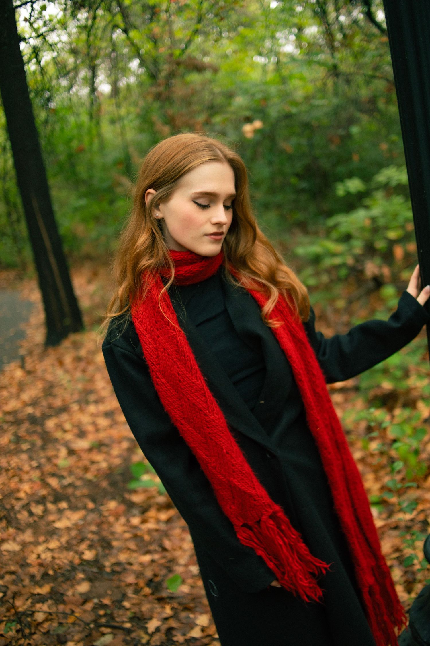 Young woman with red scarf and black coat standing in autumn forest with fallen leaves.