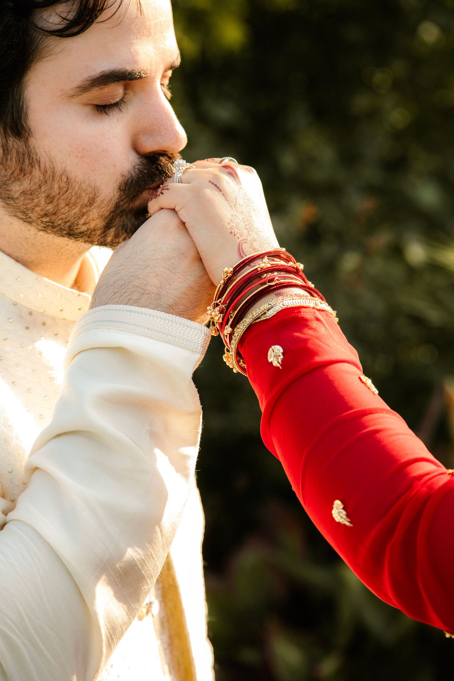 Groom in white sherwani kissing bride's hand adorned with red bangles and intricate henna at Indian wedding.