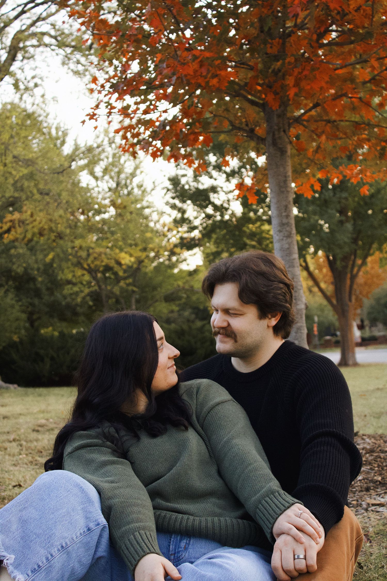 Couple sitting together on grass smiling at each other beneath vibrant autumn foliage.