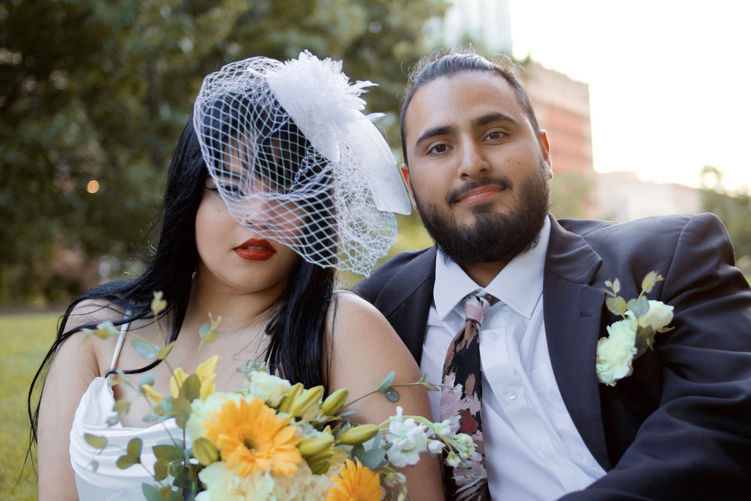 Bride in birdcage veil with red lips and groom in suit holding yellow floral bouquet outdoors.