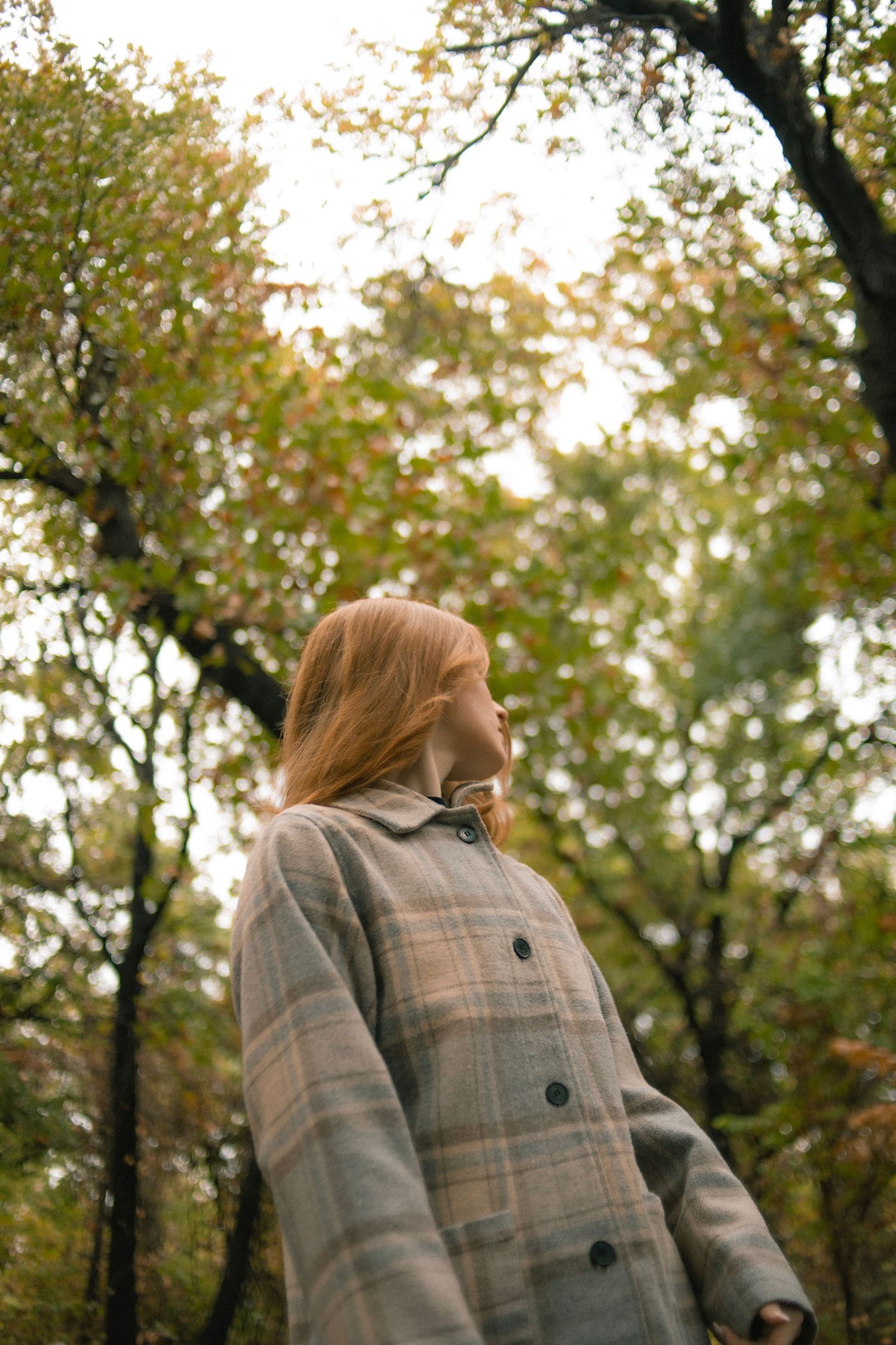 Young woman with red hair in plaid coat standing among autumn trees in a forest park.