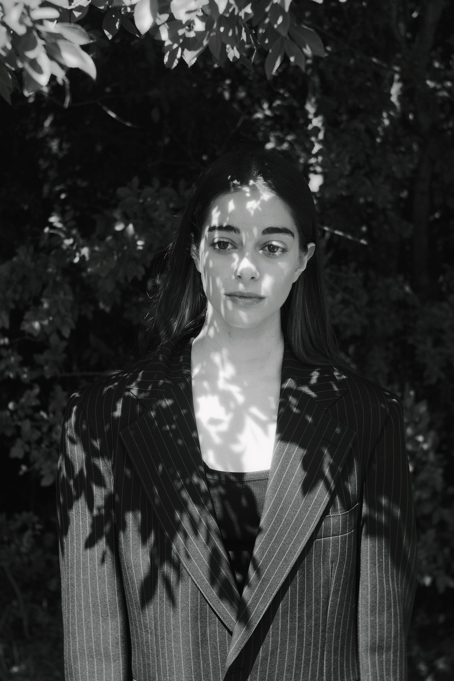 Black and white portrait of a woman in pinstripe blazer standing among trees with dappled sunlight on her face.