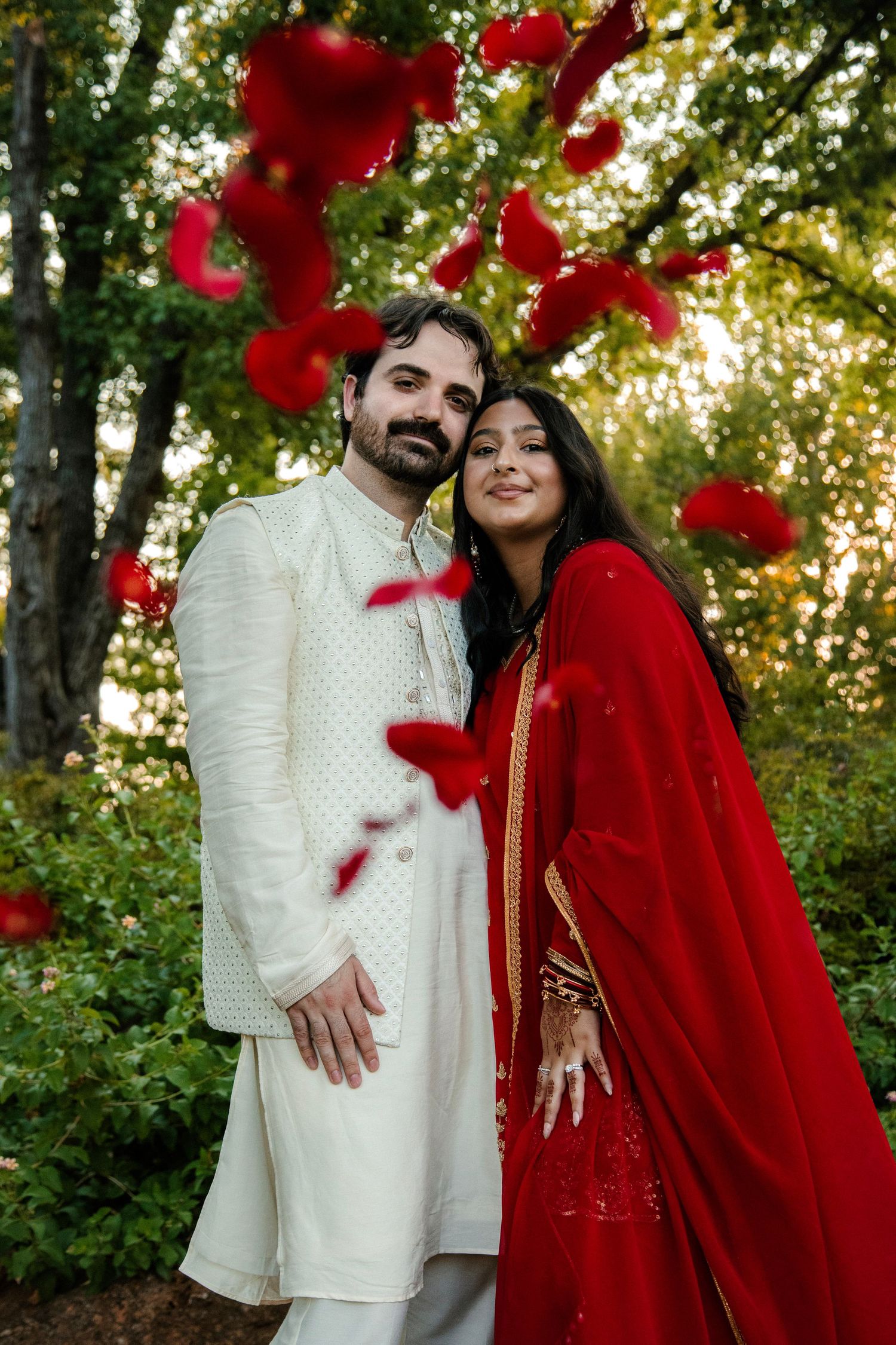 Indian wedding couple in traditional red and white attire surrounded by falling red rose petals outdoors.