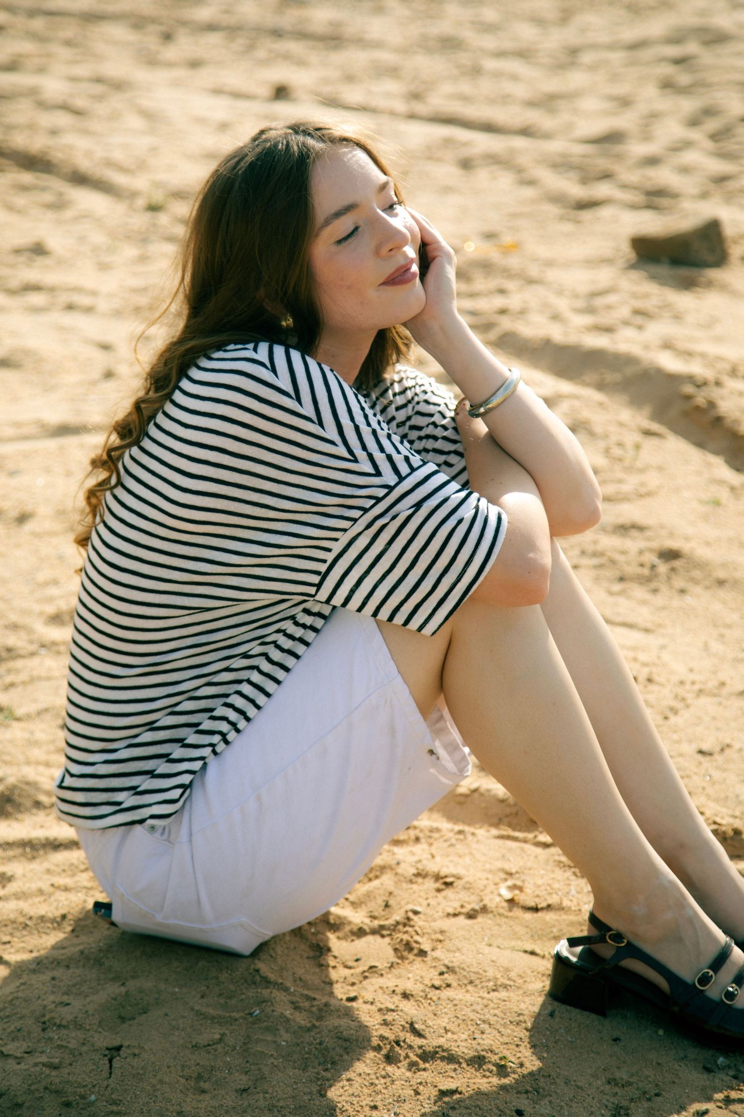 Young woman in striped top and white shorts sitting on sandy beach in warm sunlight.