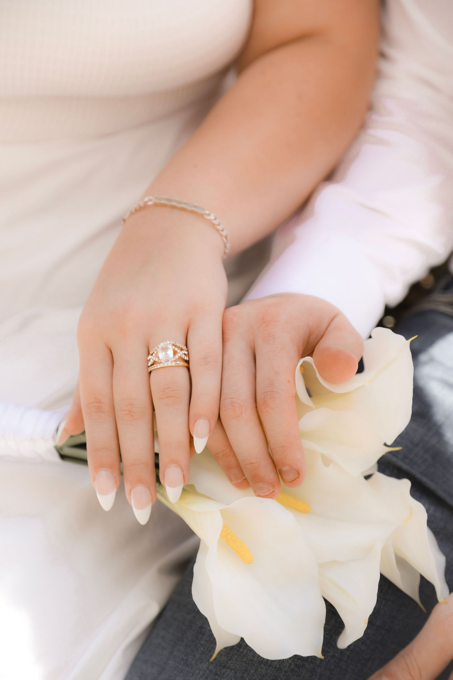 Close-up of a bride's hand with gold engagement ring and bracelet resting on white calla lilies.