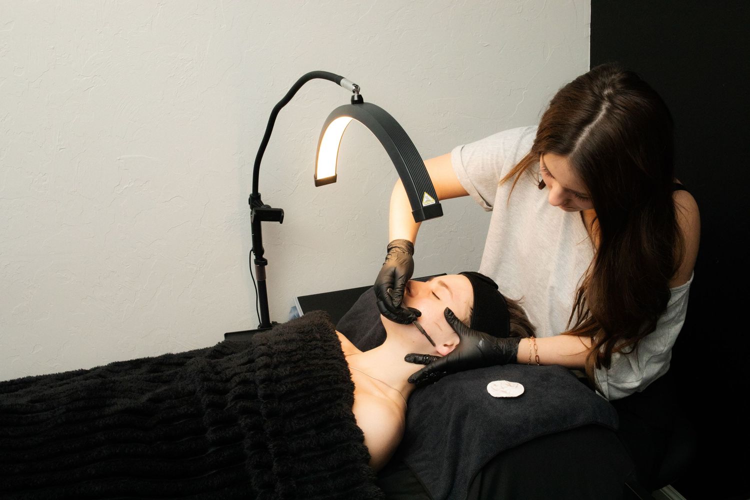 Esthetician in black gloves performing facial treatment on client under bright lamp.