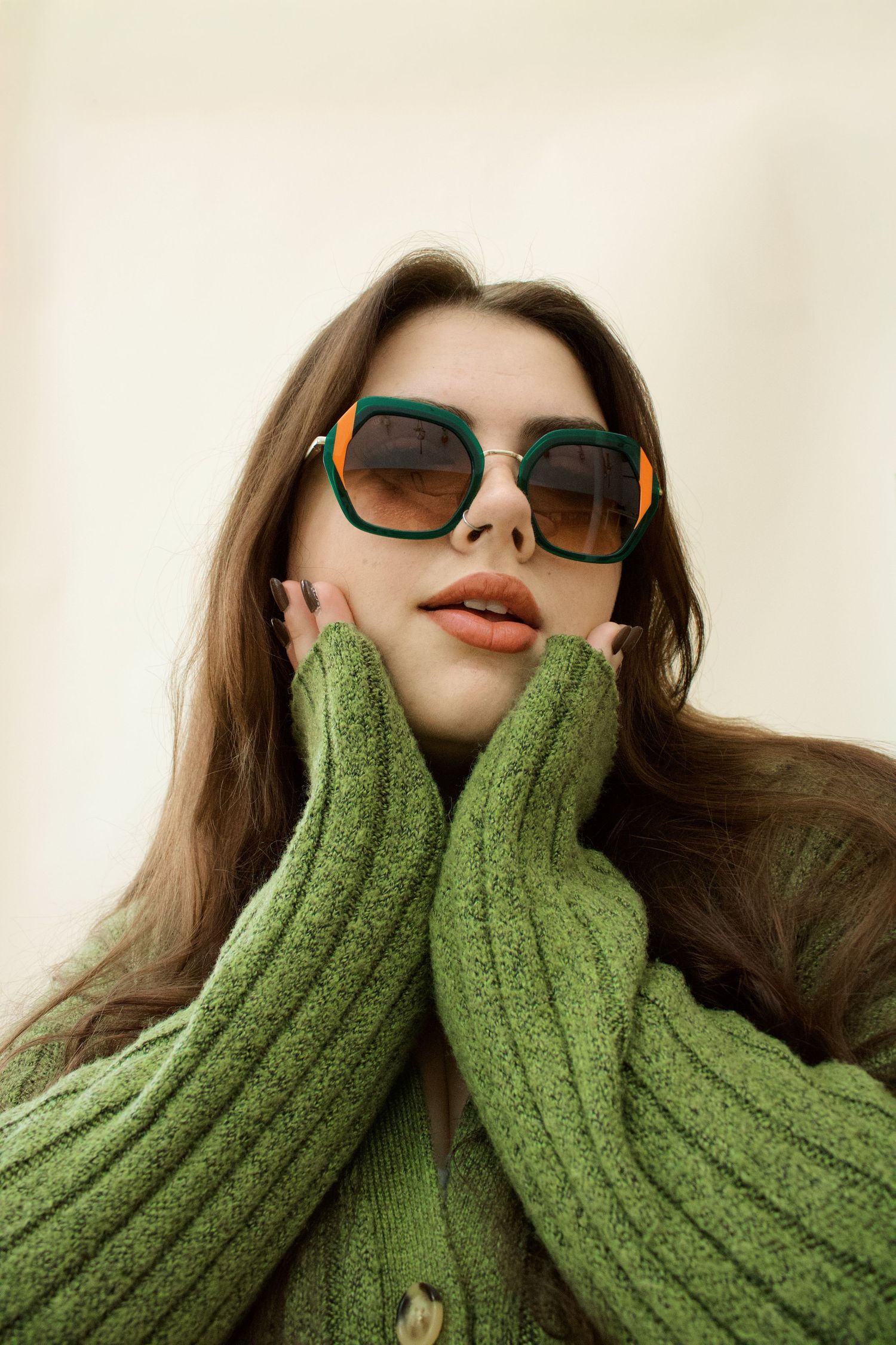 Young woman in oversized green sunglasses and green knit sweater posing with hands framing her face.