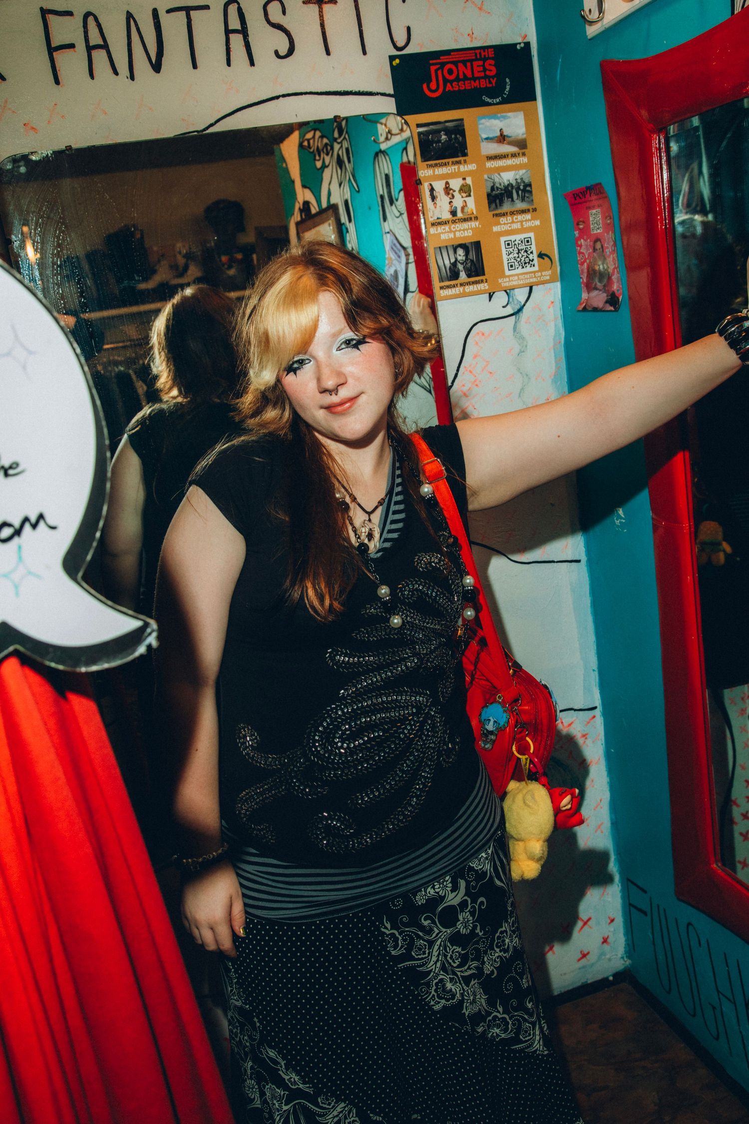 Redhead woman in black embellished top holding a red stuffed animal bag in a colorful shop.