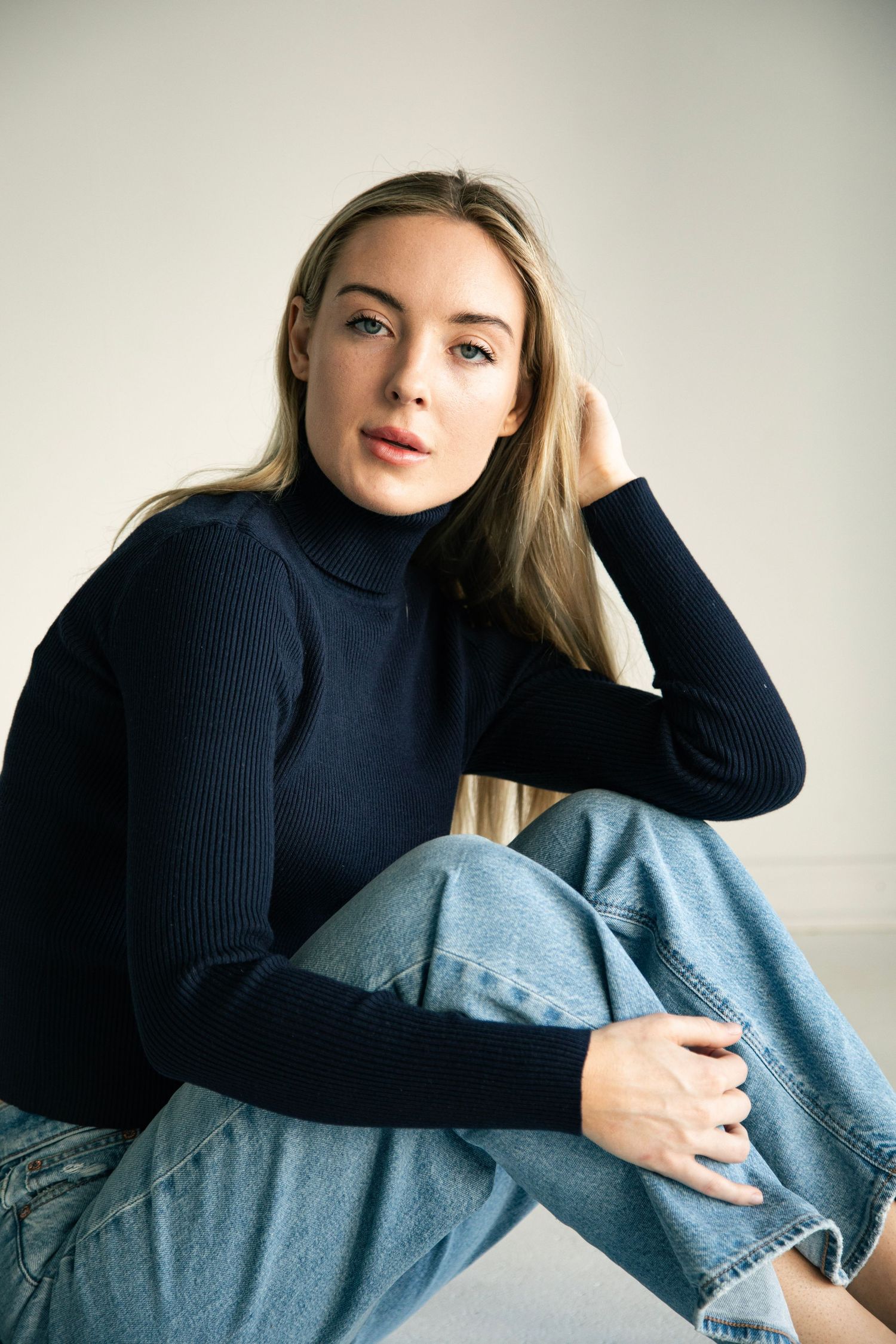 Young blonde woman in navy ribbed turtleneck and jeans sitting casually on floor.
