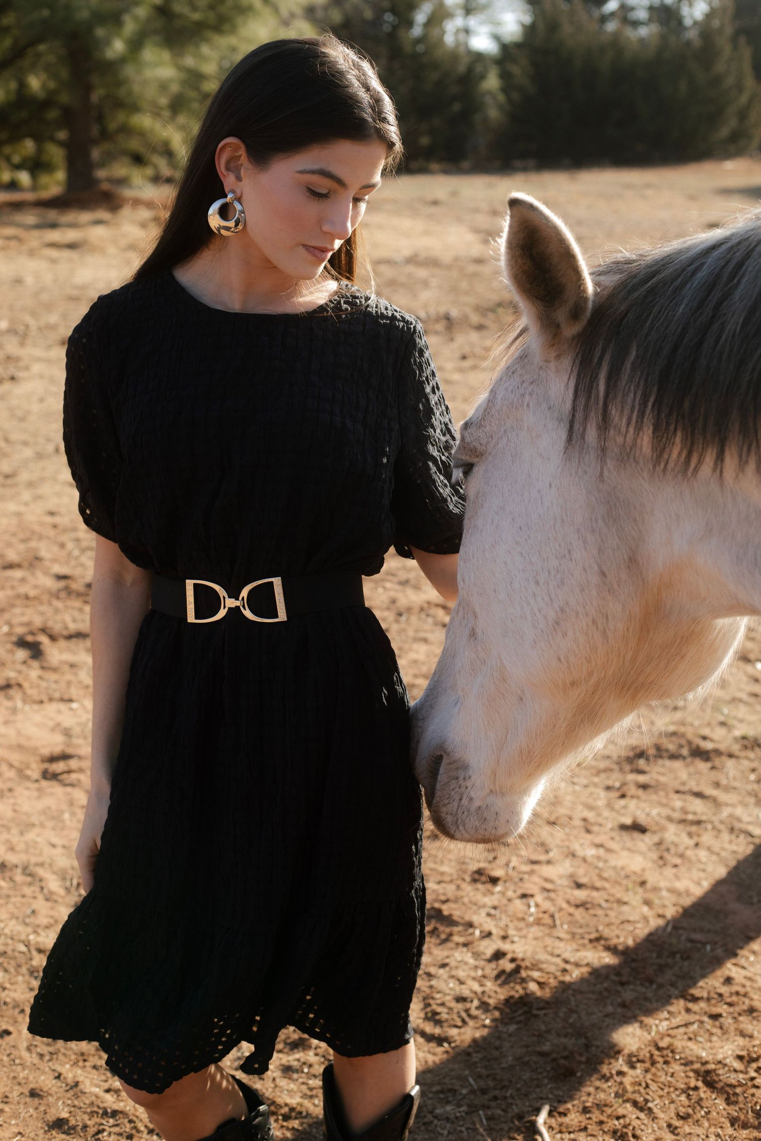 Young woman in black knit dress with gold D-ring belt standing beside white horse outdoors.