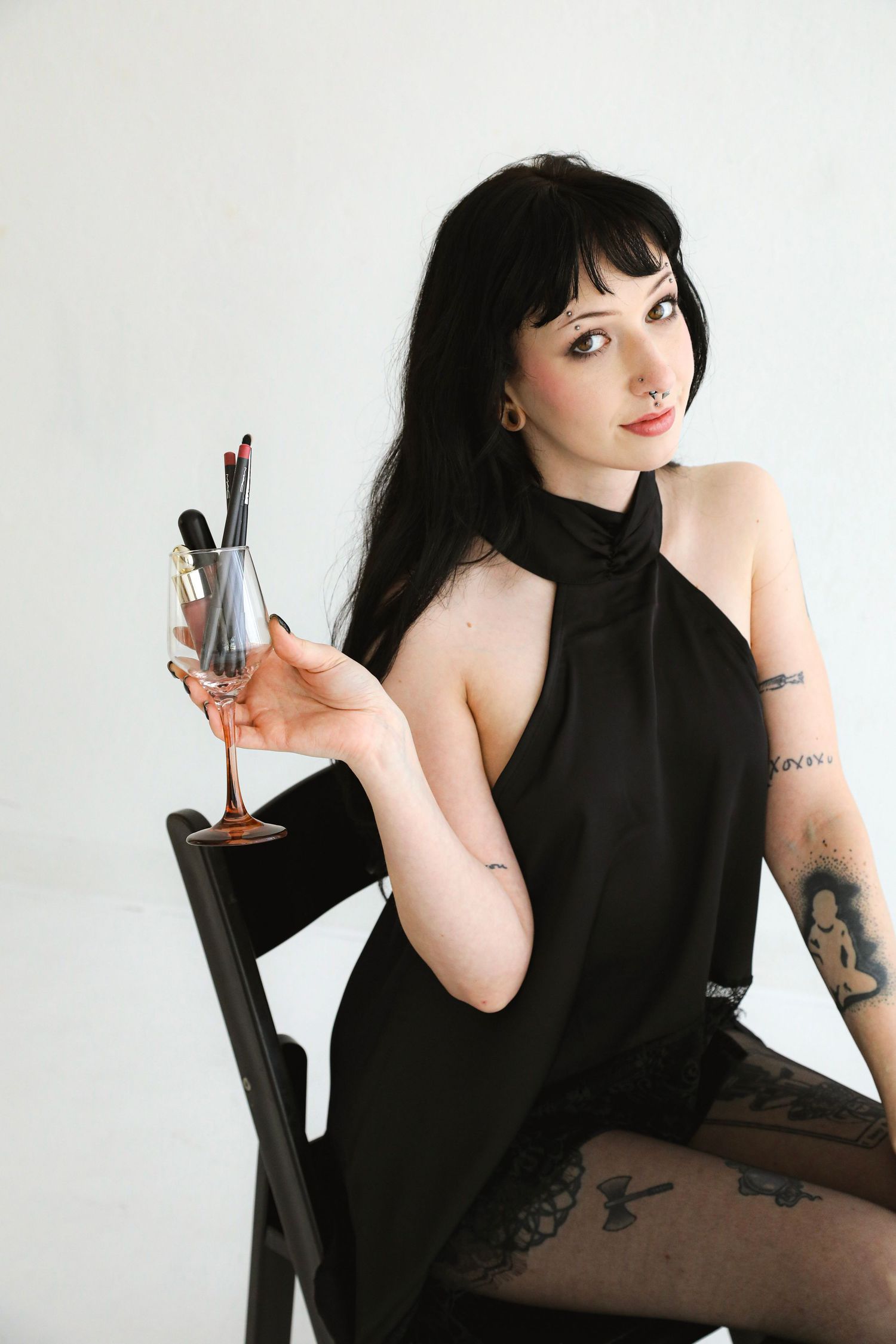 A tattooed woman in a black halter dress sits on a chair holding a wine glass with makeup brushes against a white background.