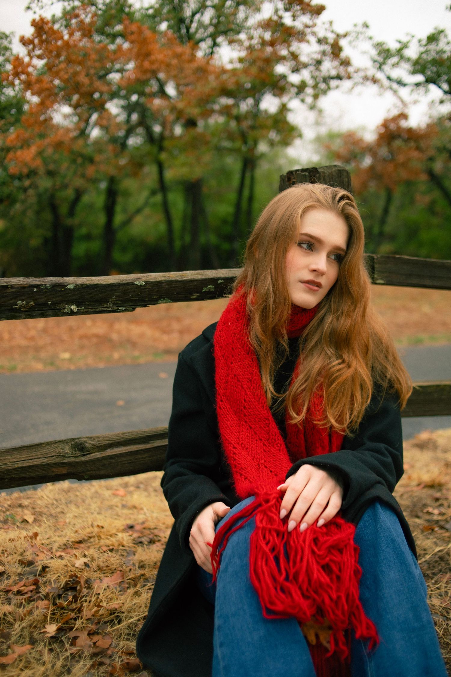 Young woman with long red hair wearing a black coat and red scarf sits on a wooden fence in an autumn park.
