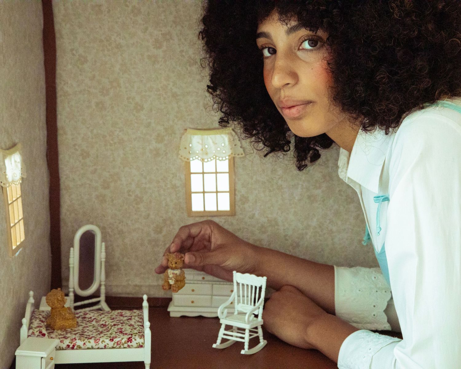 Woman with natural curly hair interacts with a detailed miniature dollhouse bedroom scene.