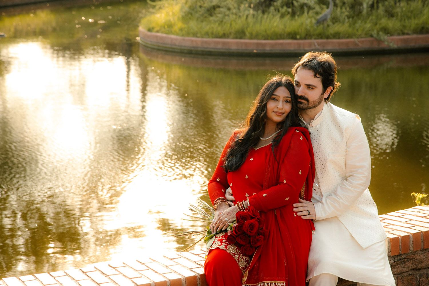 Indian bride and groom in red and white traditional wedding attire sitting together by a golden reflective pond.