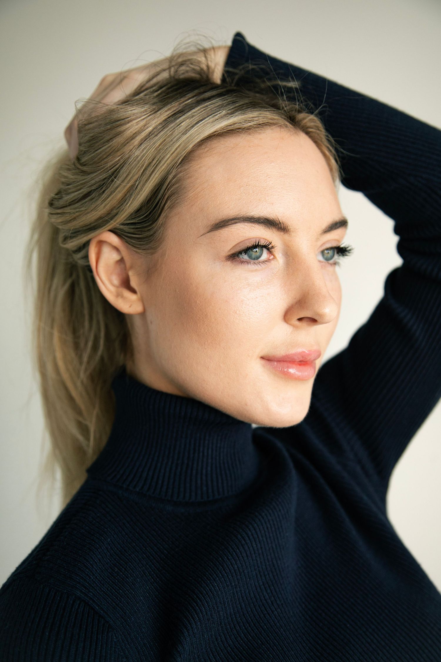 Blonde woman in black turtleneck sweater posing with hand behind head against white background.