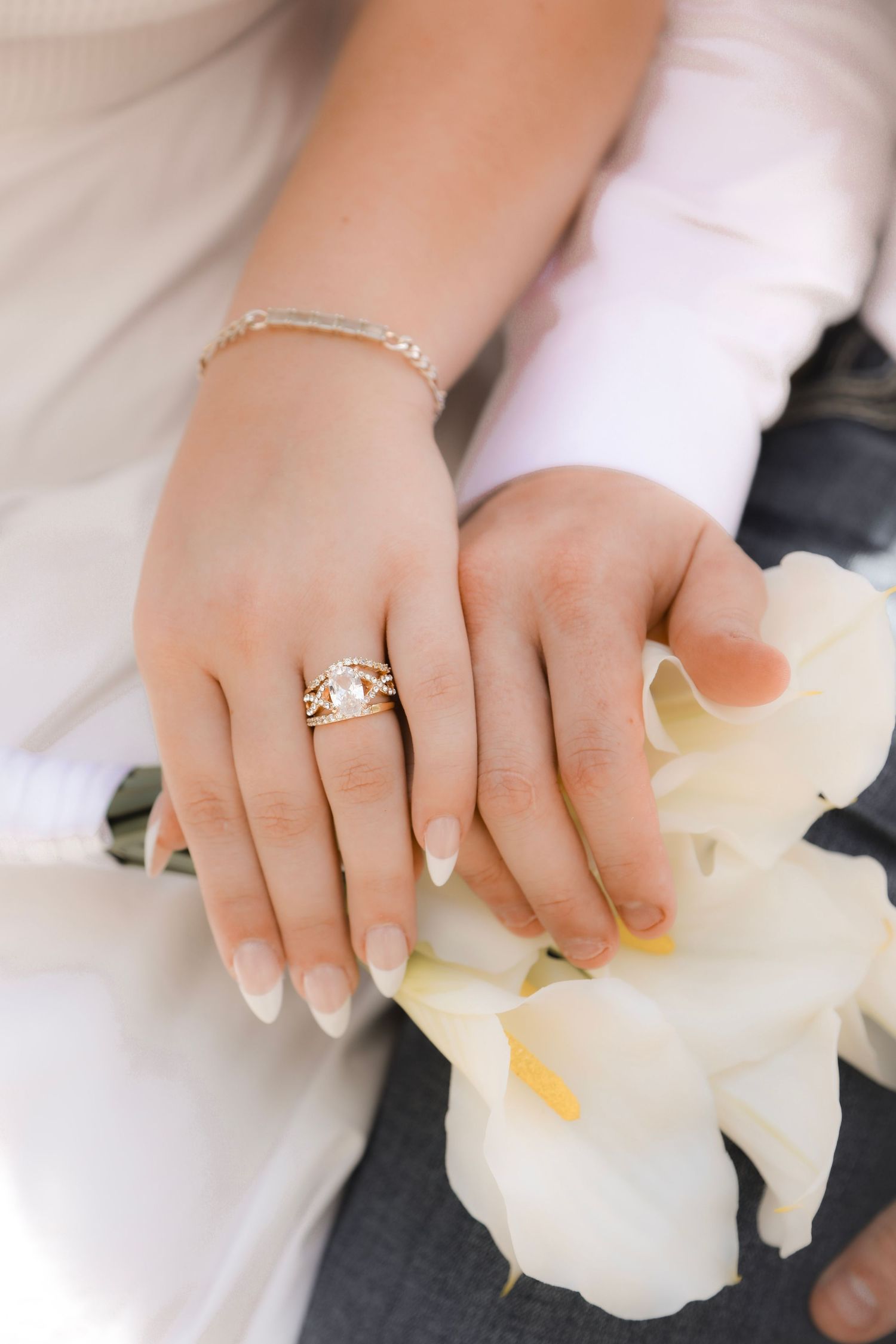 Close-up of bride's hands wearing pearl gold ring and bracelet resting on white calla lilies.