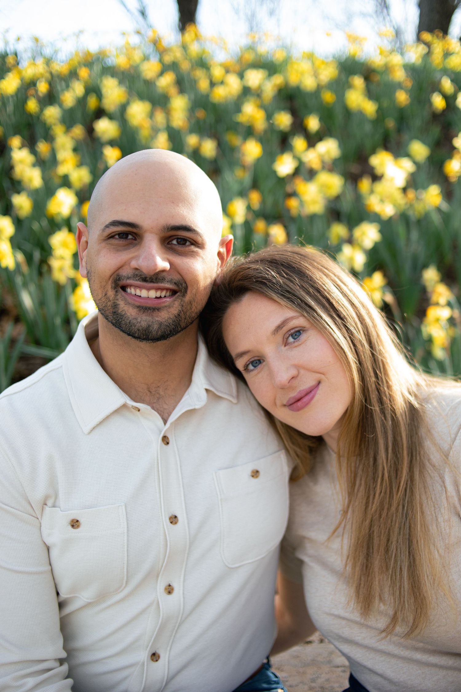 Happy engaged couple posing together in front of bright yellow daffodil flowers outdoors.