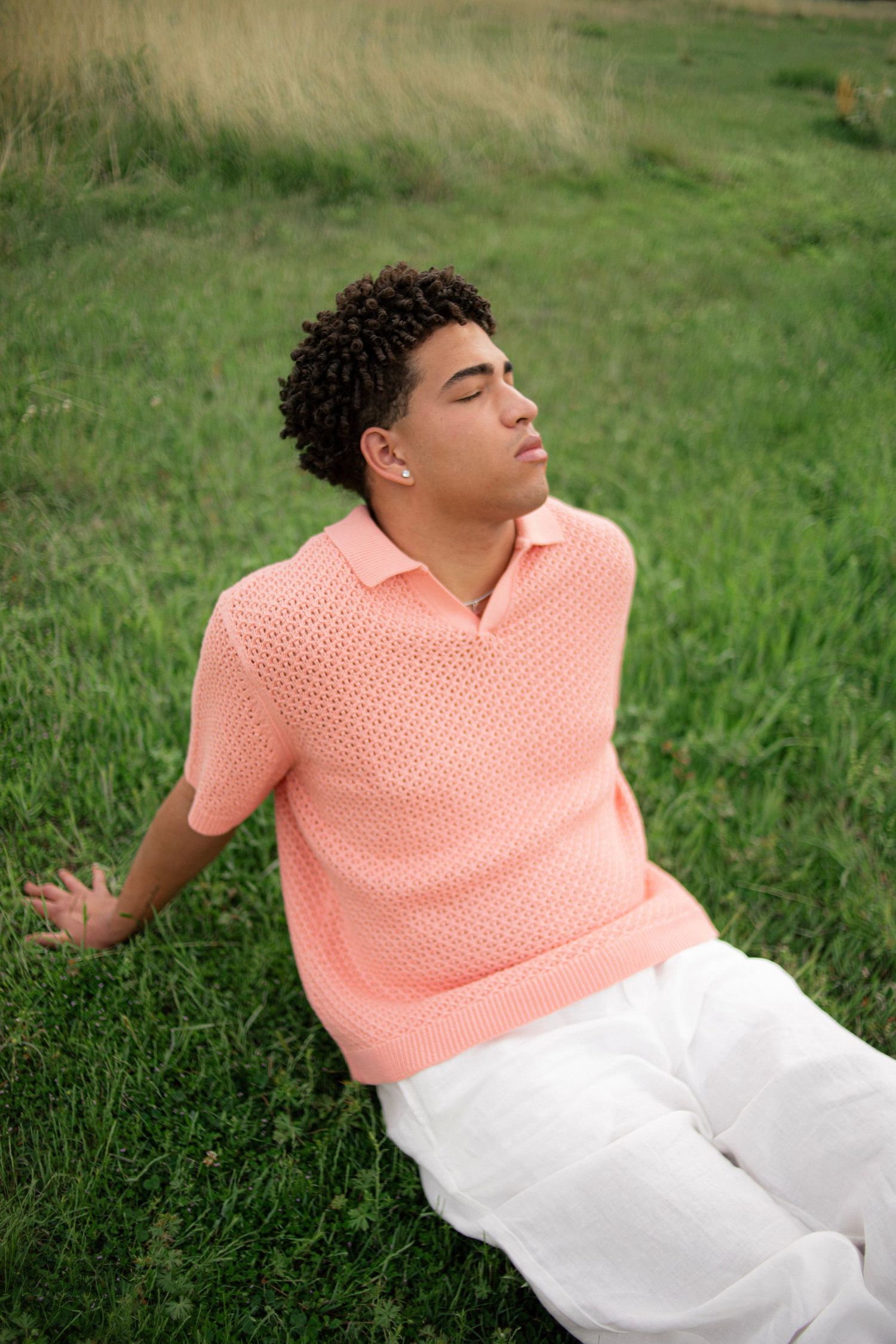 Young man in pink knit polo and white pants relaxing on green grass with eyes closed outdoors.