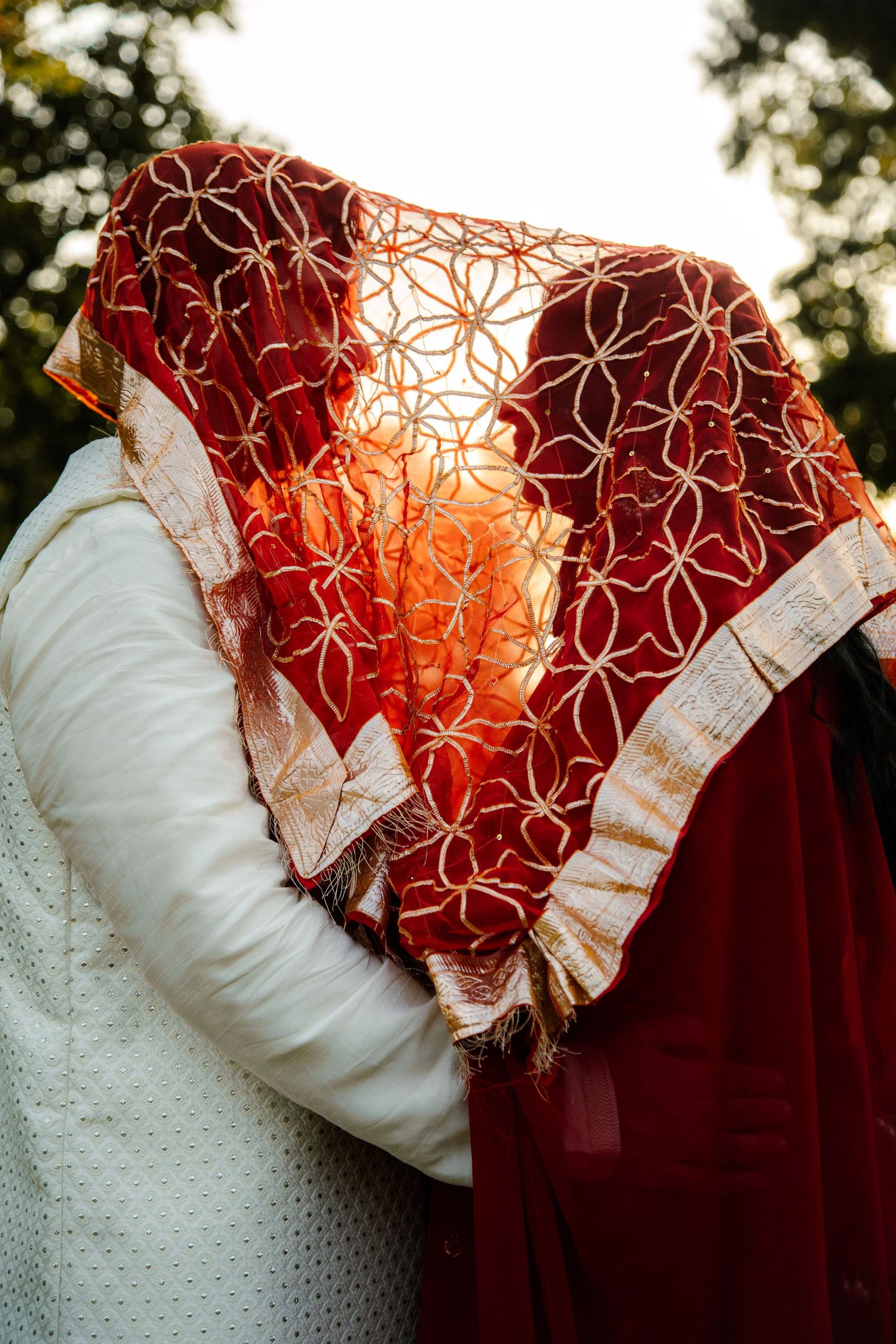 Indian wedding couple sharing a kiss hidden beneath an ornate red embroidered dupatta in golden sunlight.