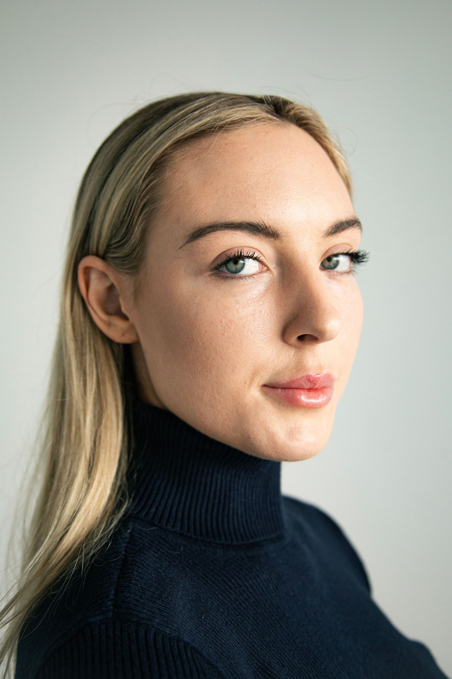 Portrait of a blonde woman in a dark navy turtleneck sweater against a light gray background.