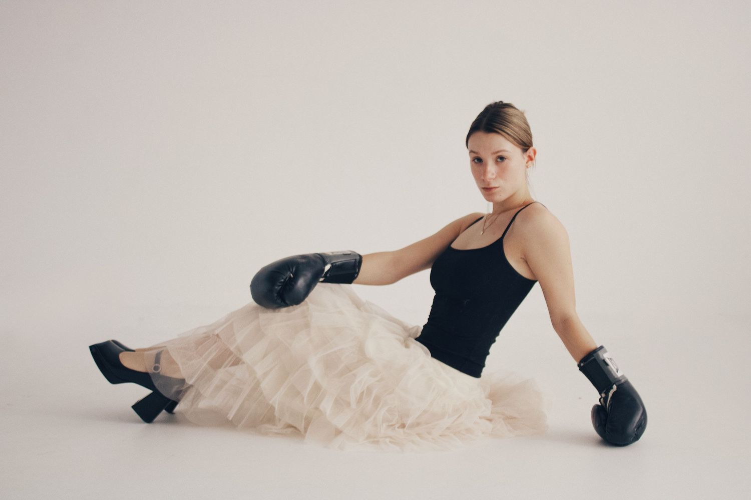 Woman in black bodysuit, tulle skirt, and boxing gloves sitting gracefully on white floor.