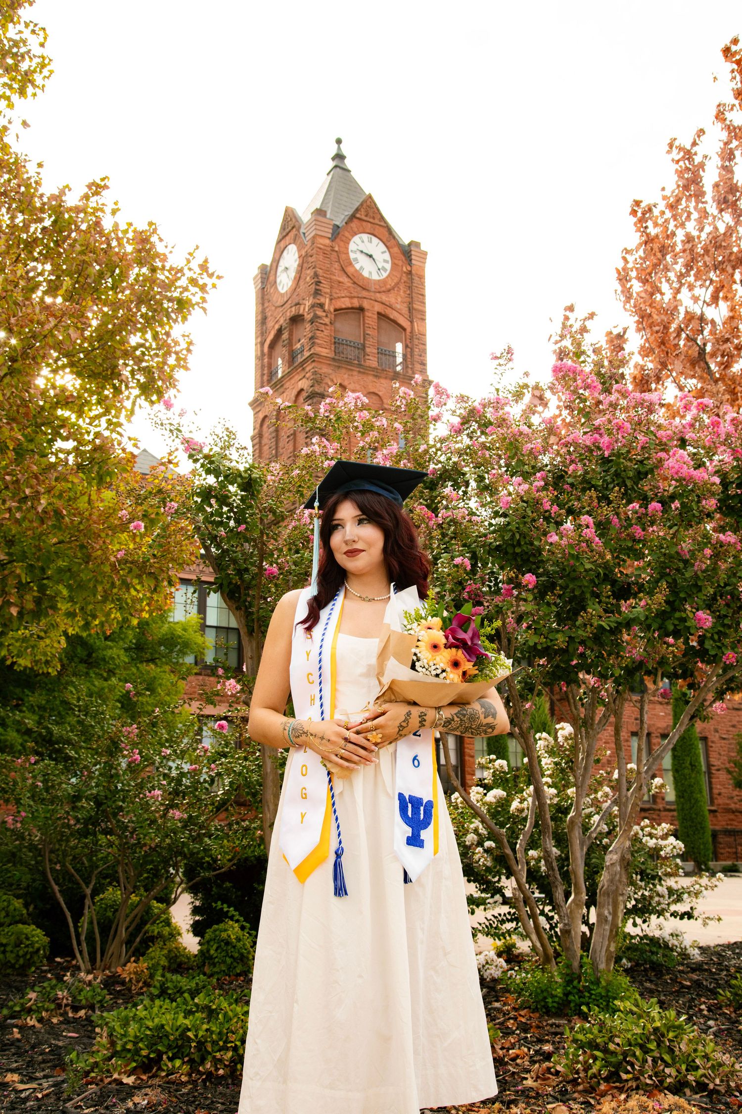 Young woman in white graduation gown and cap holds flowers near a red brick clock tower on a college campus.