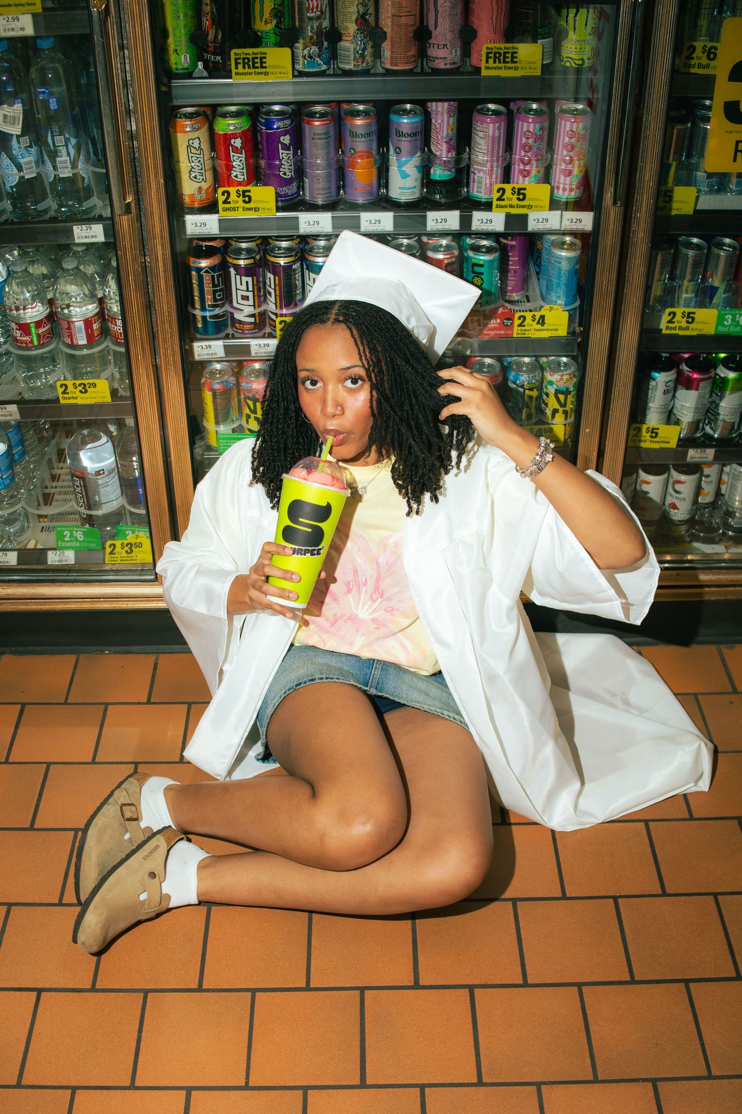 Young woman in white graduation cap and gown sitting on store floor, sipping a drink near colorful beverage coolers.
