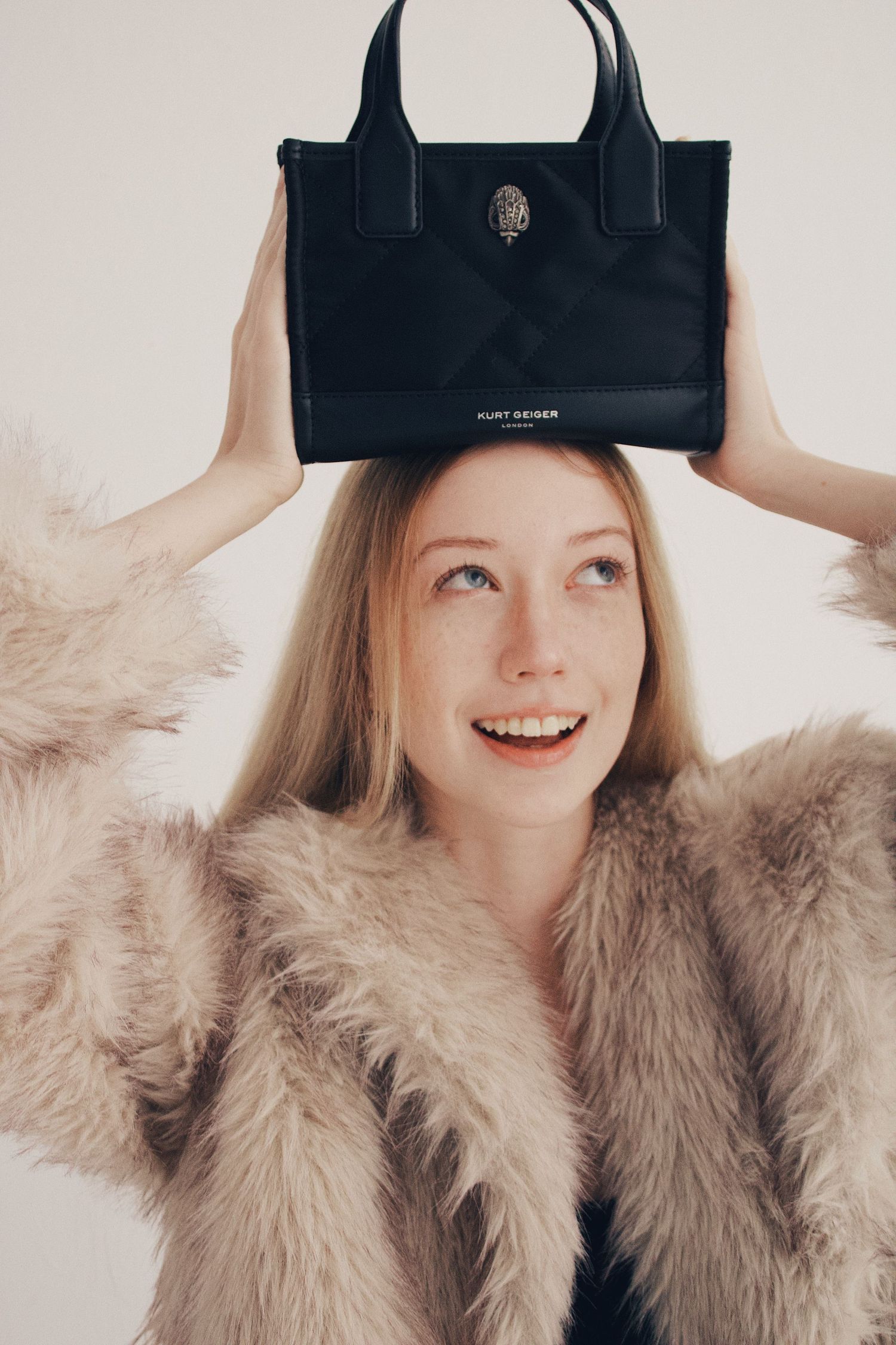Smiling blonde woman in fluffy beige coat balancing black Kurt Geiger tote bag on her head.