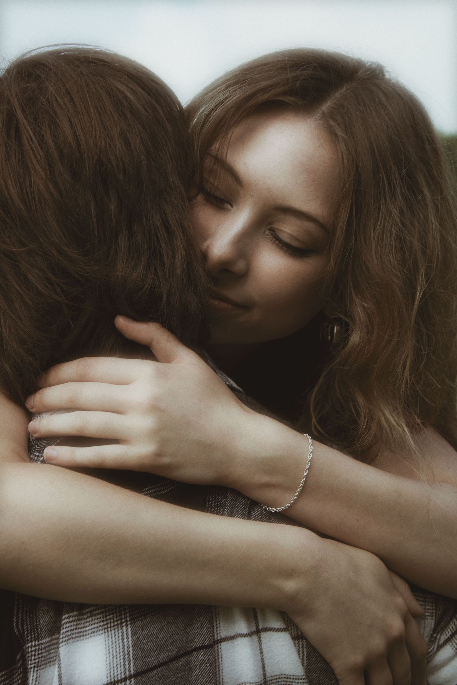 A young woman with brown hair tenderly embraces someone outdoors, eyes closed, wearing a silver bracelet.