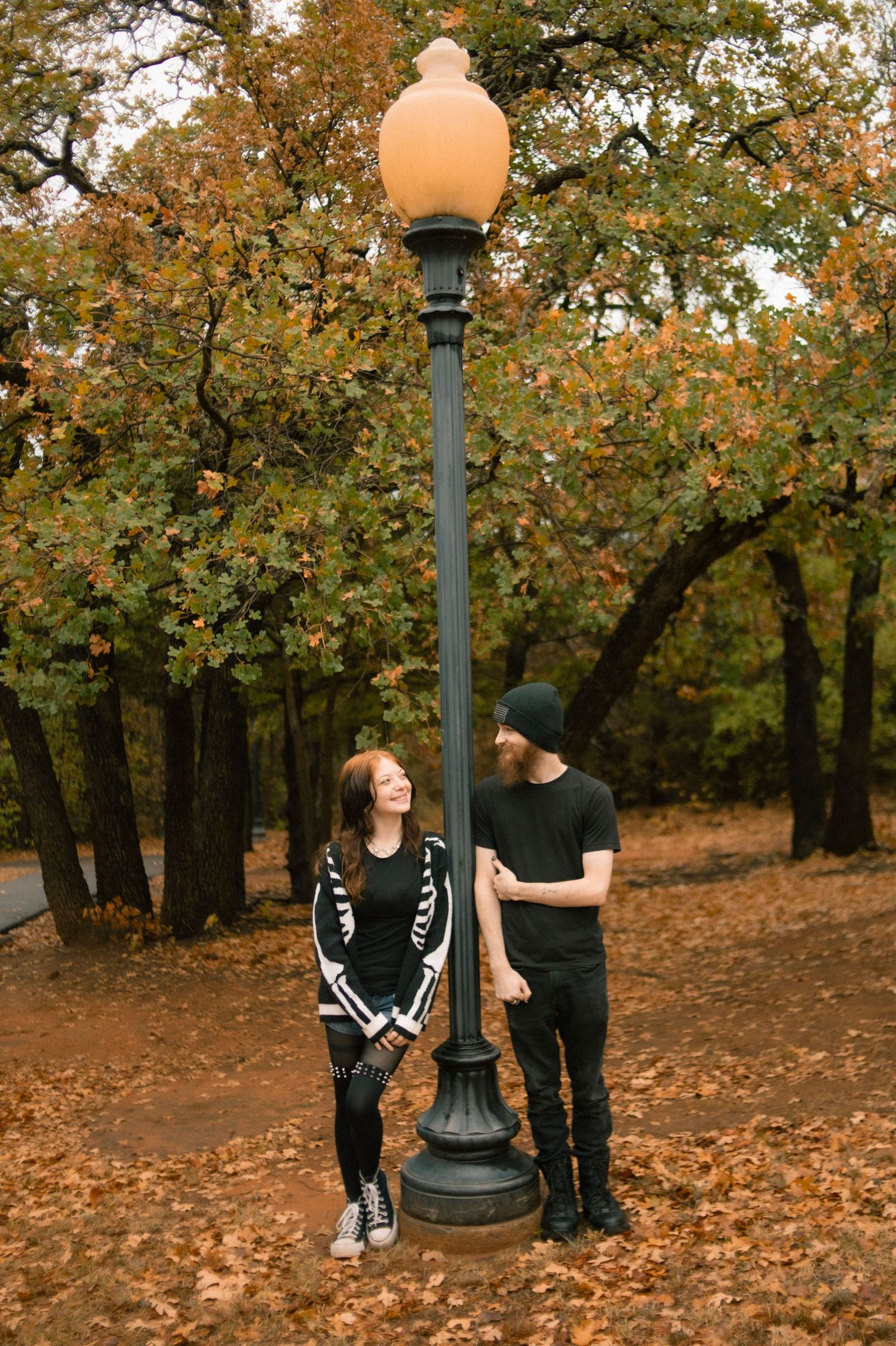 A couple stands by a vintage street lamp in an autumn park, surrounded by fallen leaves and colorful foliage.