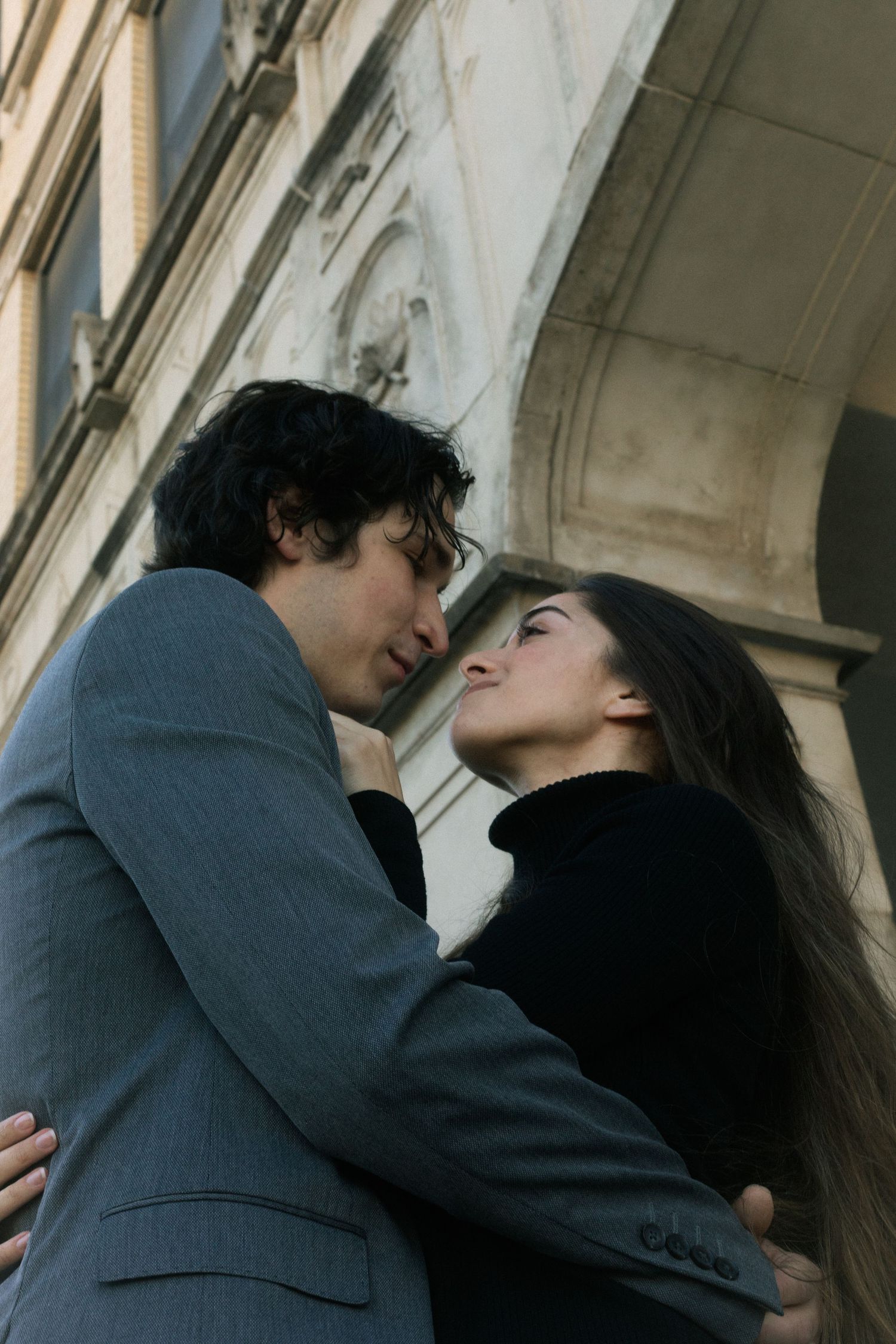 A romantic couple gazes into each other's eyes while embracing near a classic stone building arch.