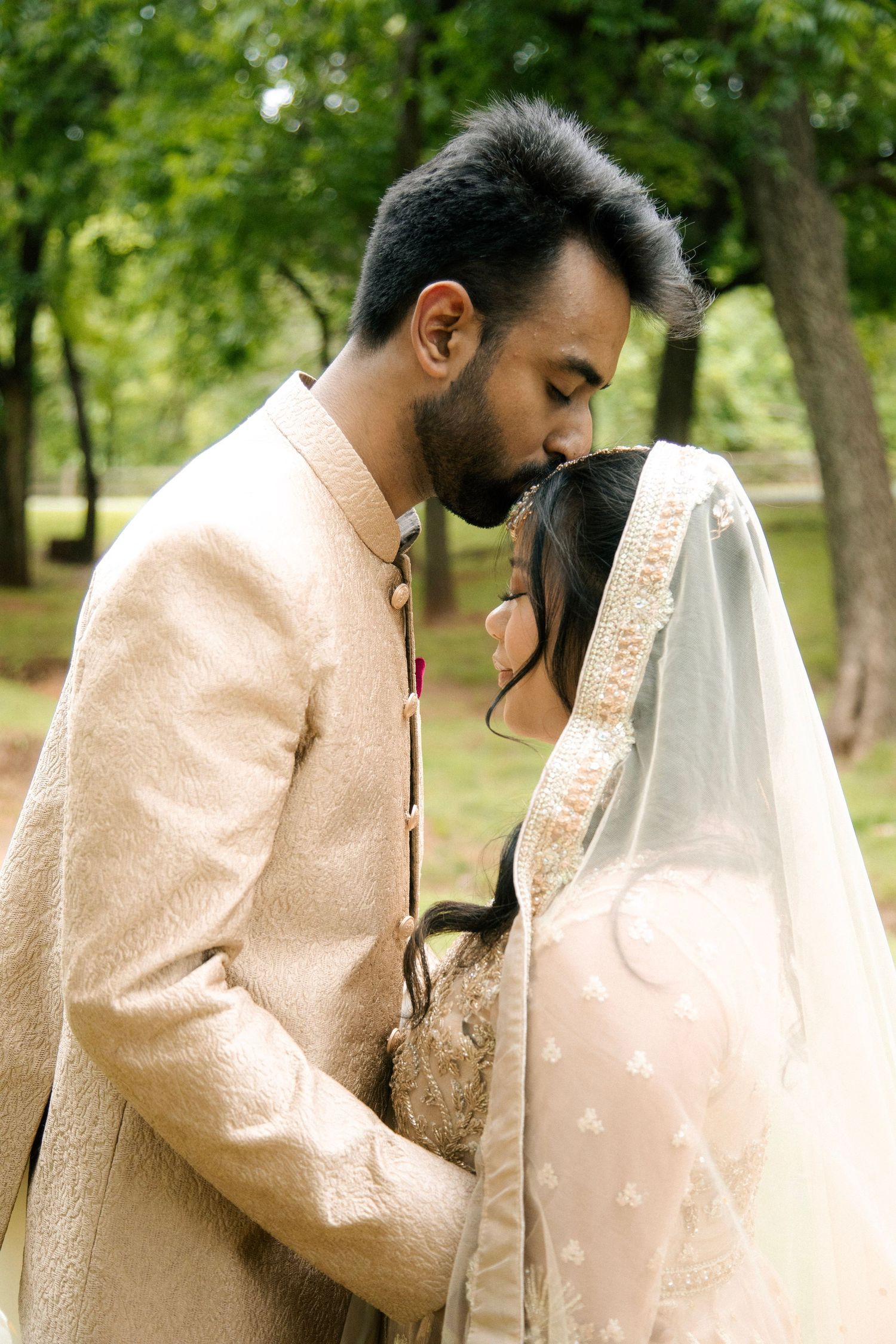 Groom in cream sherwani tenderly kissing bride's forehead as she wears a delicate veil in a lush green park.