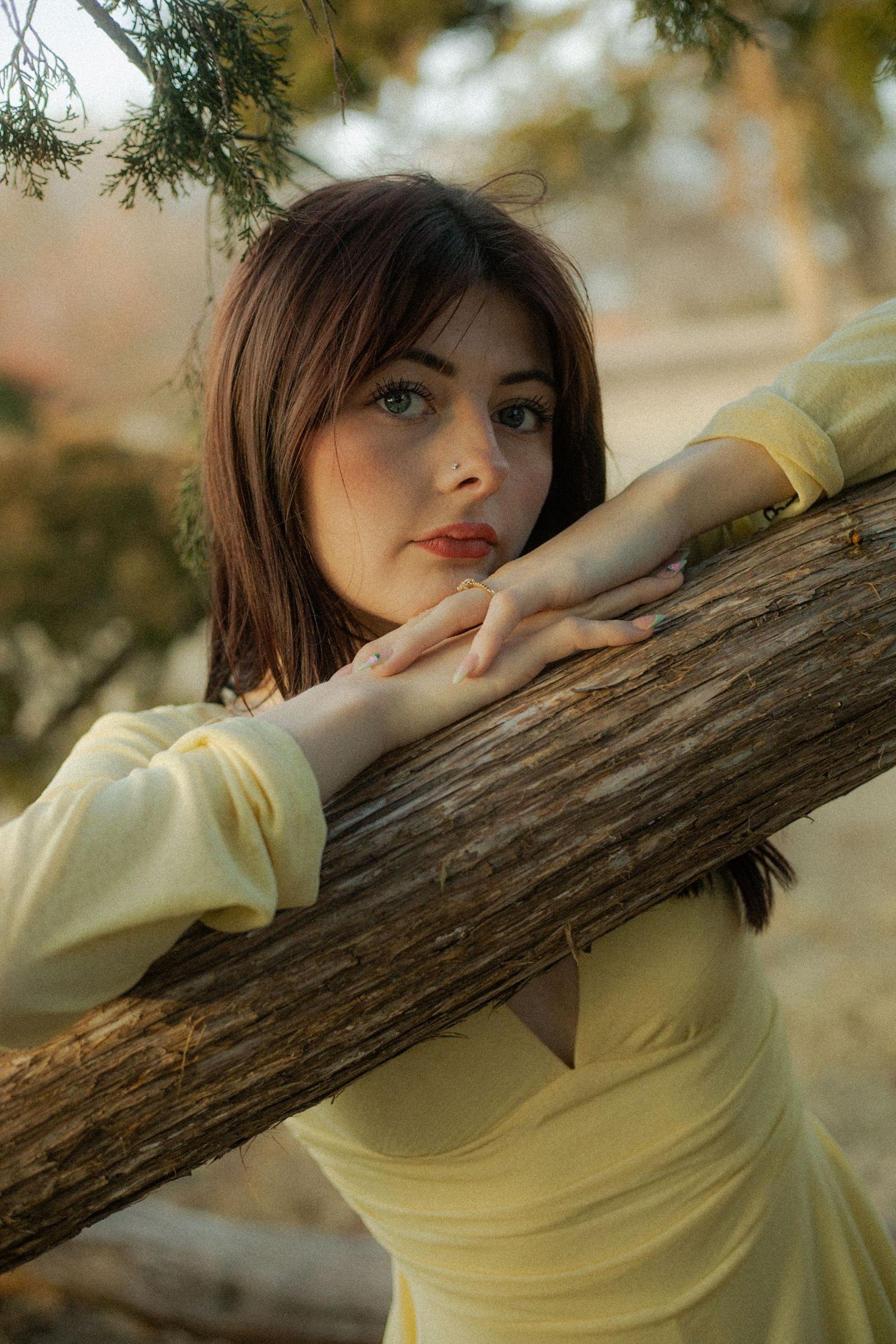 Young woman with blue eyes in yellow dress leaning on rustic wooden log surrounded by trees.