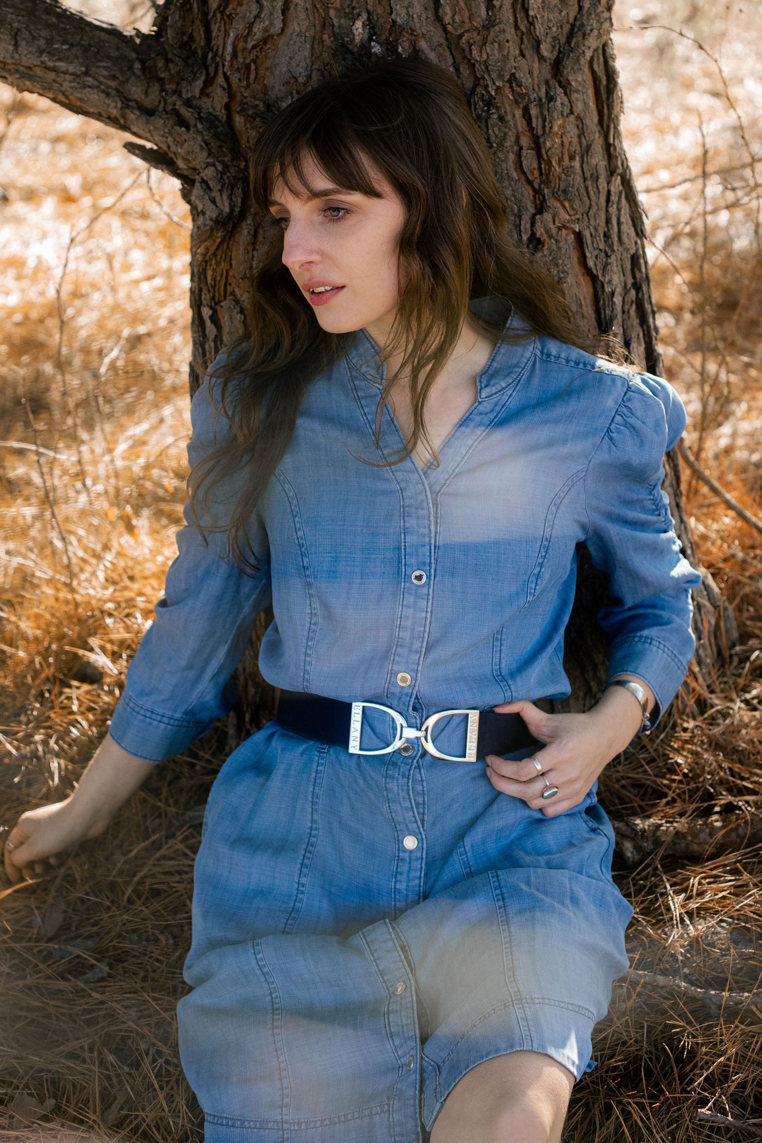 Woman in blue denim dress with black belt leaning against tree in golden field.