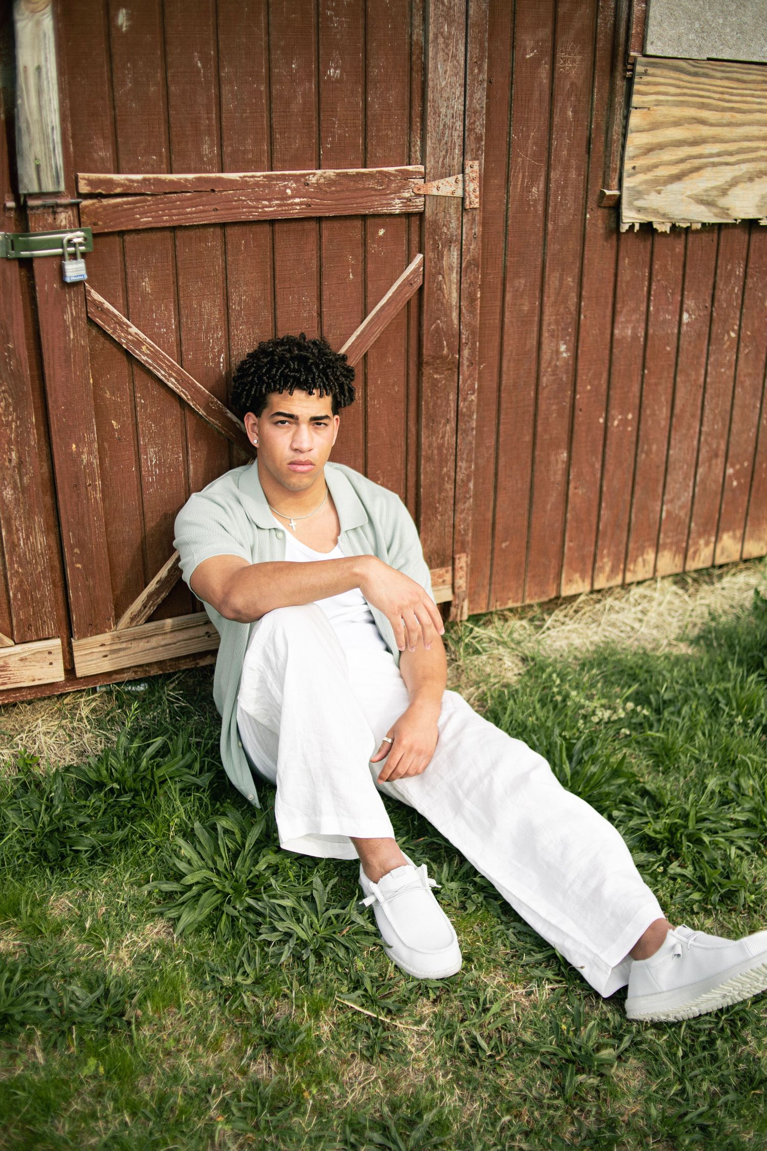 Young man in white pants and sage shirt sitting against rustic wooden barn door on green grass.