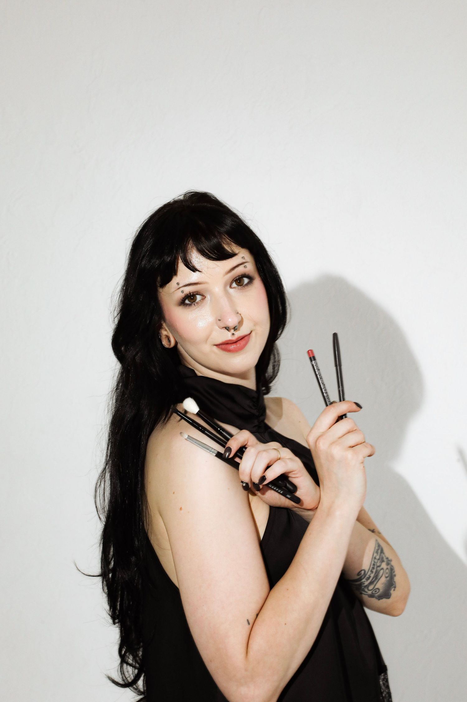 Dark-haired makeup artist holding brushes and lipstick, posing confidently with a tattoo visible on her arm.