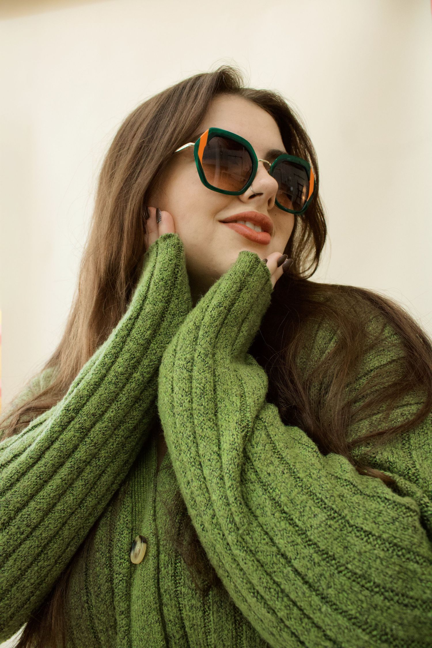 Brunette woman in oversized green knit sweater and green sunglasses posing against light background.