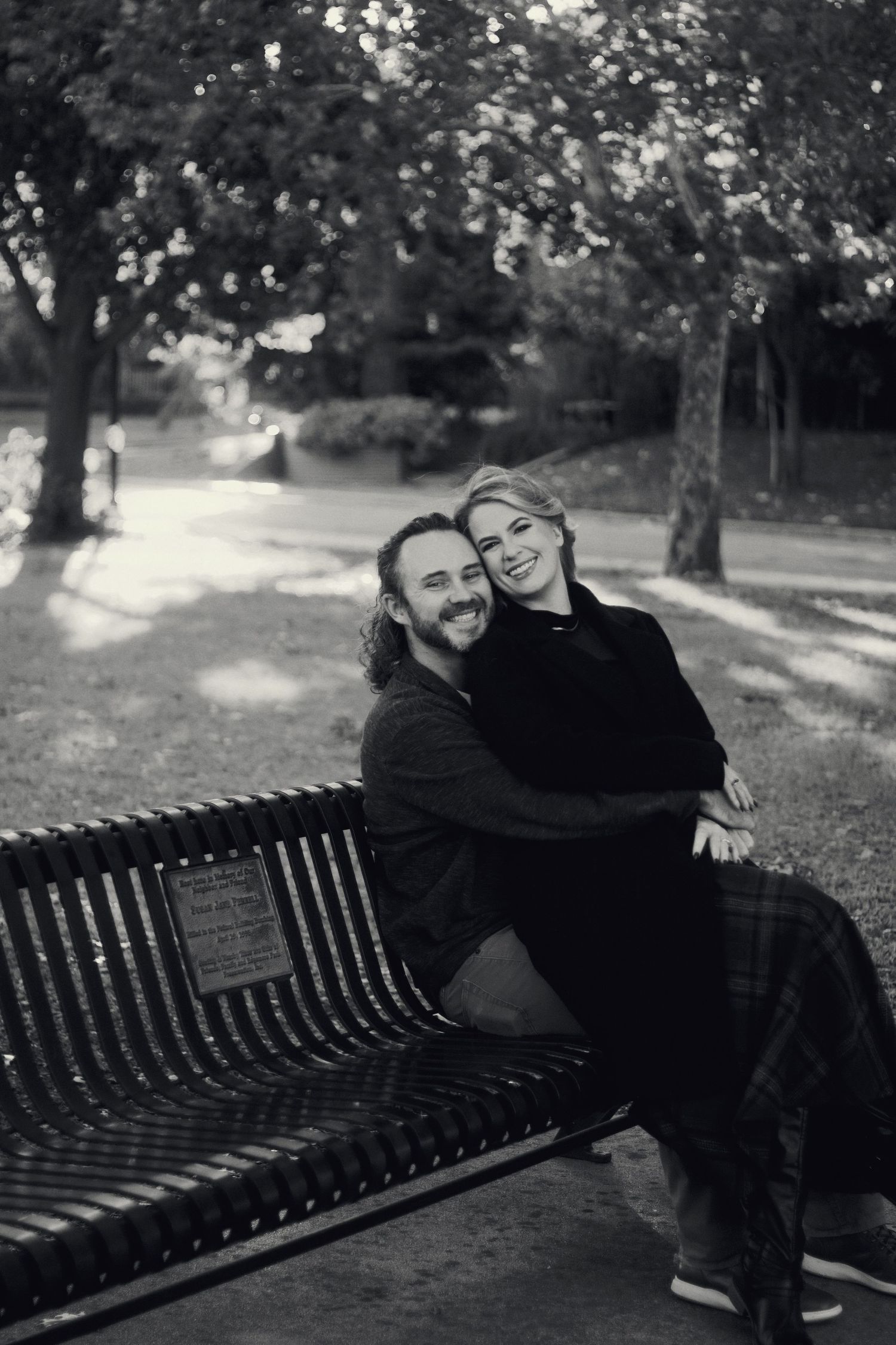 Black and white photo of smiling couple embracing on a park bench surrounded by trees.