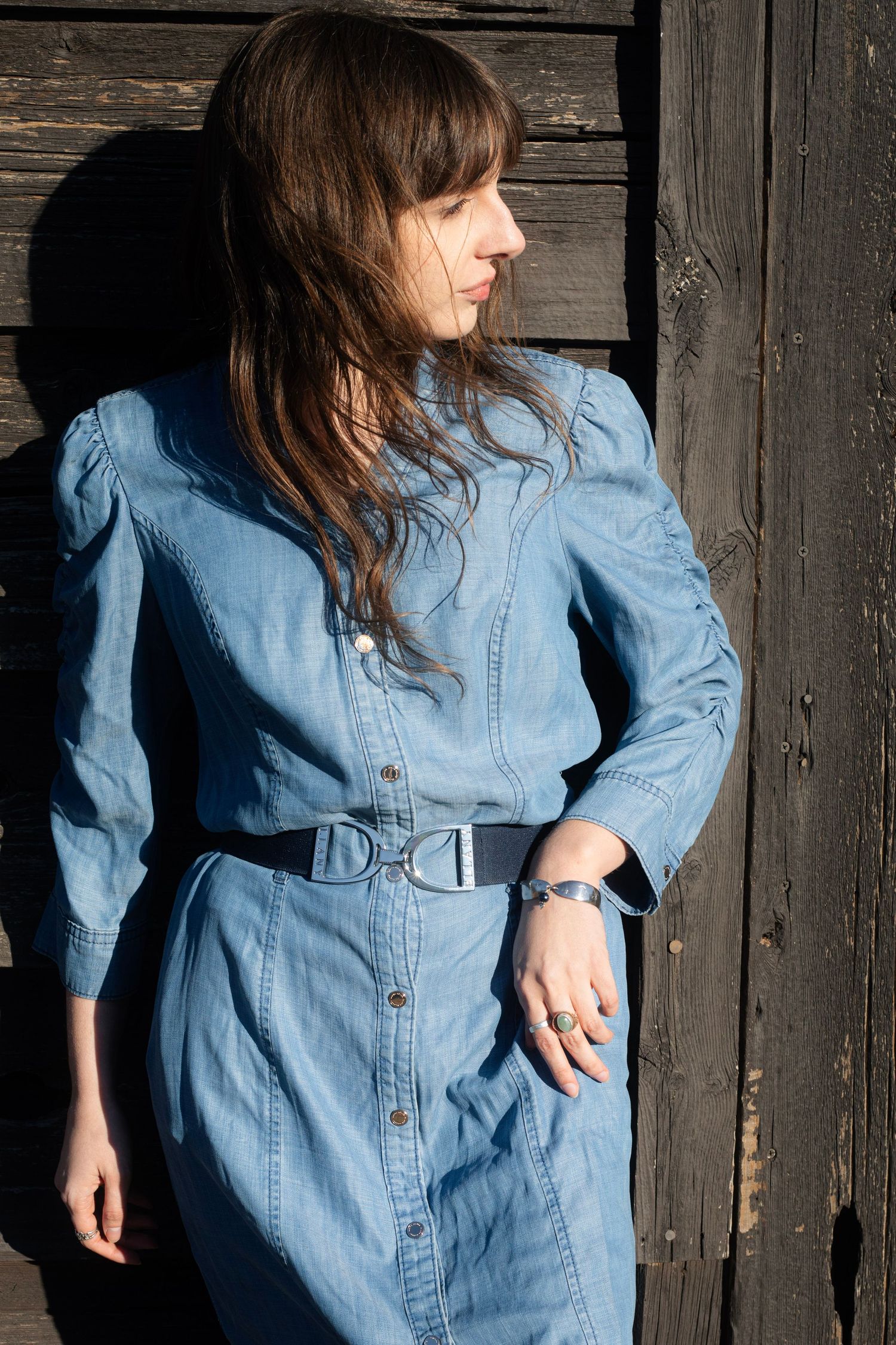 Woman in blue denim button-down dress with belt leaning against rustic dark wood wall in sunlight.