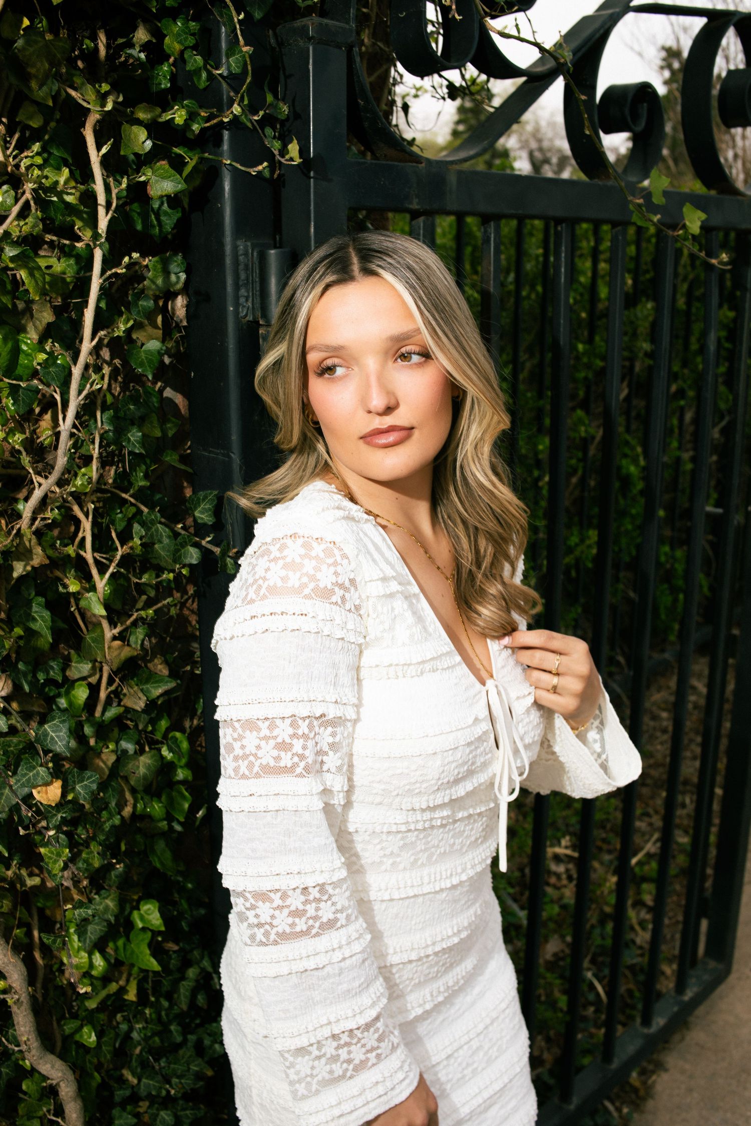 Young woman in white lace dress posing by black iron gate with ivy.